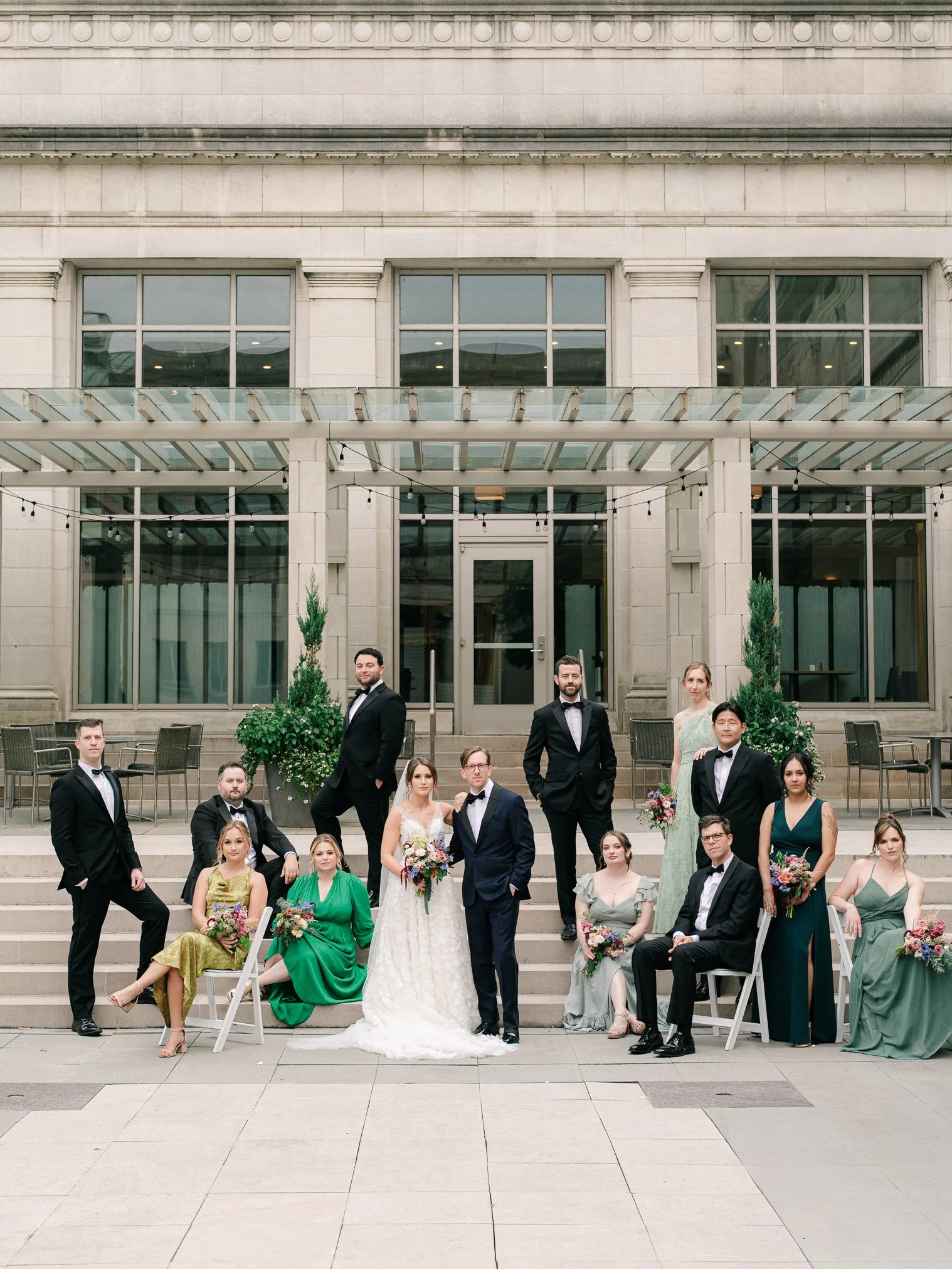 A group of people in formal attire on steps outside a building, including a bride in a white wedding dress and a groom in a suit, surrounded by bridesmaids and groomsmen, with some holding bouquets of flowers.