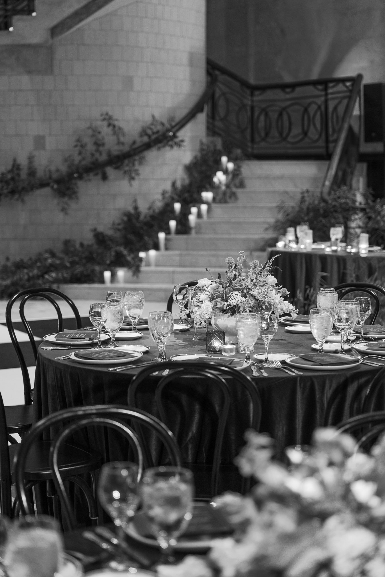 Elegant banquet table setup with floral centerpiece, water glasses, plates, and candles, in a dimly lit venue with staircase in the background.