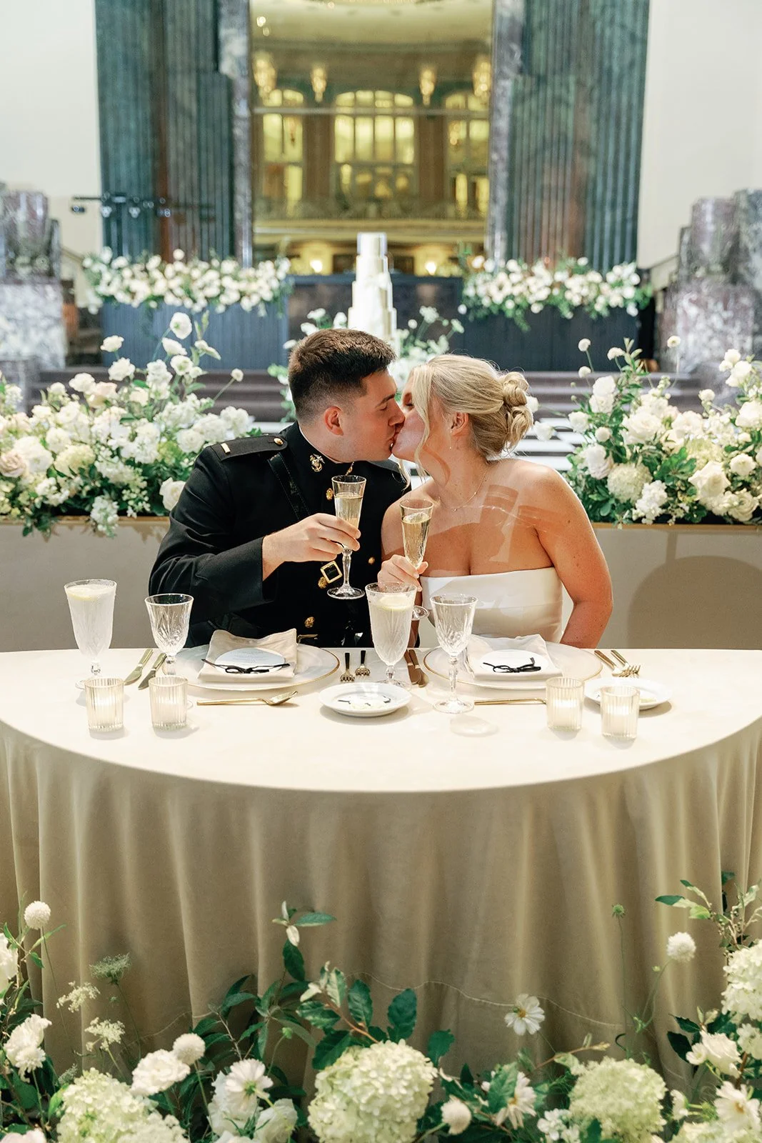 Hall of Mirrors Wedding Photography. A couple in wedding attire kissing at a sweetheart table decorated with white flowers, with champagne flutes in hand, in a grand hall with ornate architecture in the background.