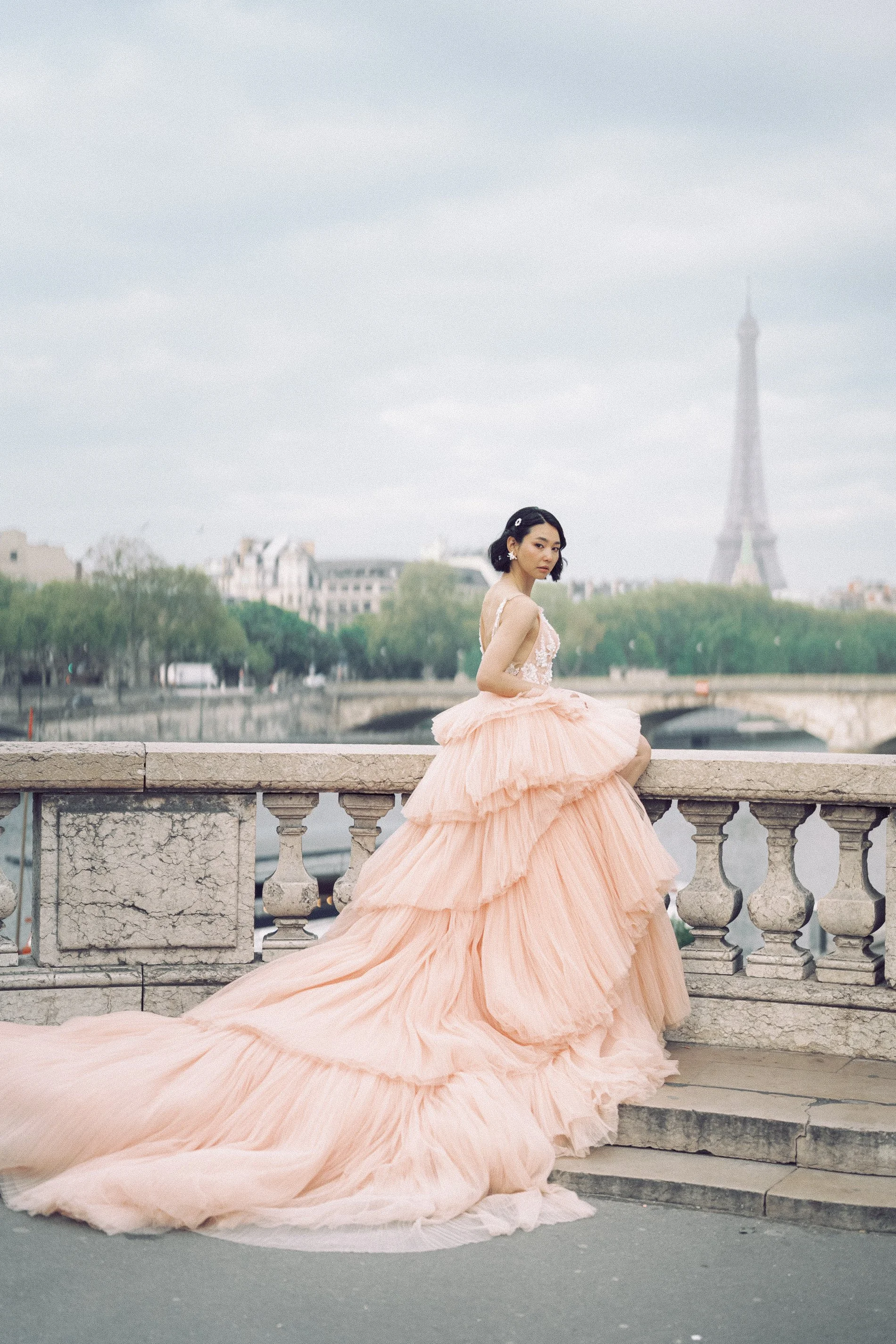 A woman in a peach ball gown sitting on a stone railing near the Seine River in Paris, with the Eiffel Tower in the background.