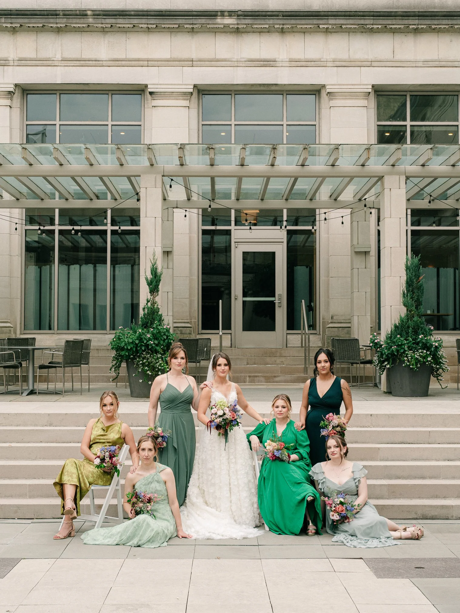 A group of seven women dressed in formal gowns, holding bouquets, poses on steps outside a building with large windows and potted plants.