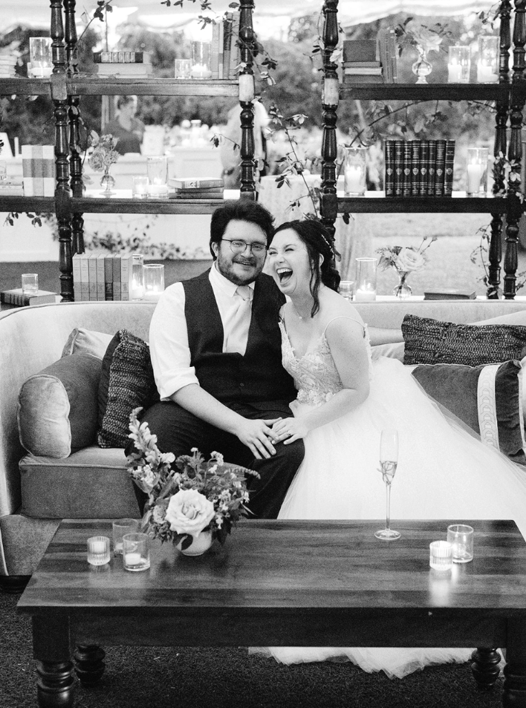 A wedding celebration with a smiling bride and groom sitting on a sofa, surrounded by candles and floral arrangements, in an elegant indoor setting.