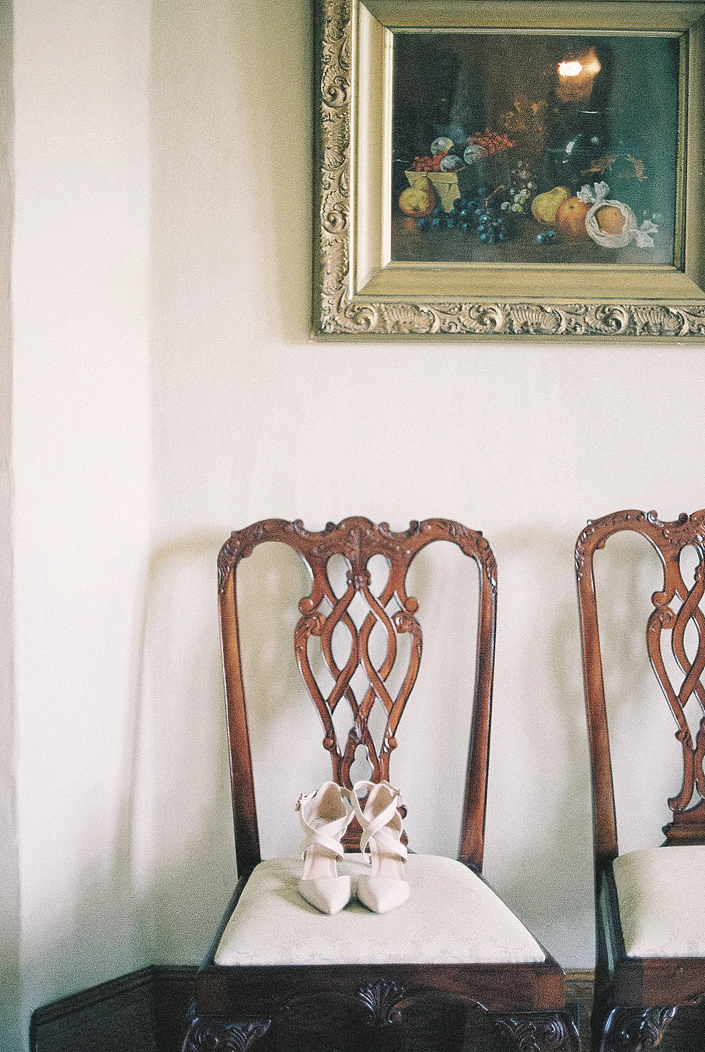 A pair of white high-heeled shoes with ankle straps placed on a carved wooden chair, with another similar chair beside it, in a room with a framed still life painting of fruit on the wall.