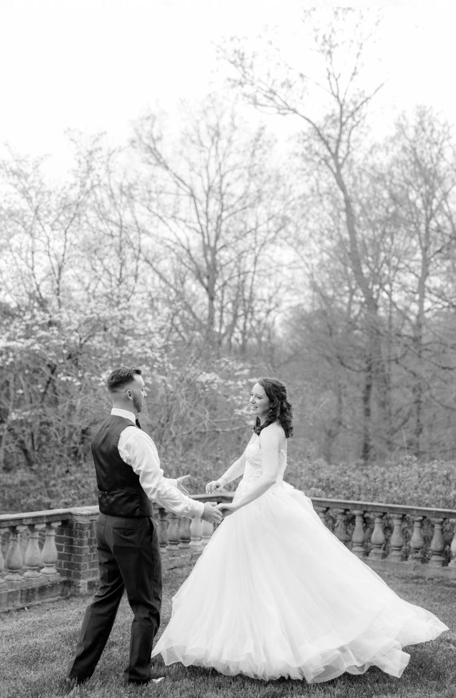 Bride and groom dancing outdoors near a railing, surrounded by trees, in black and white.