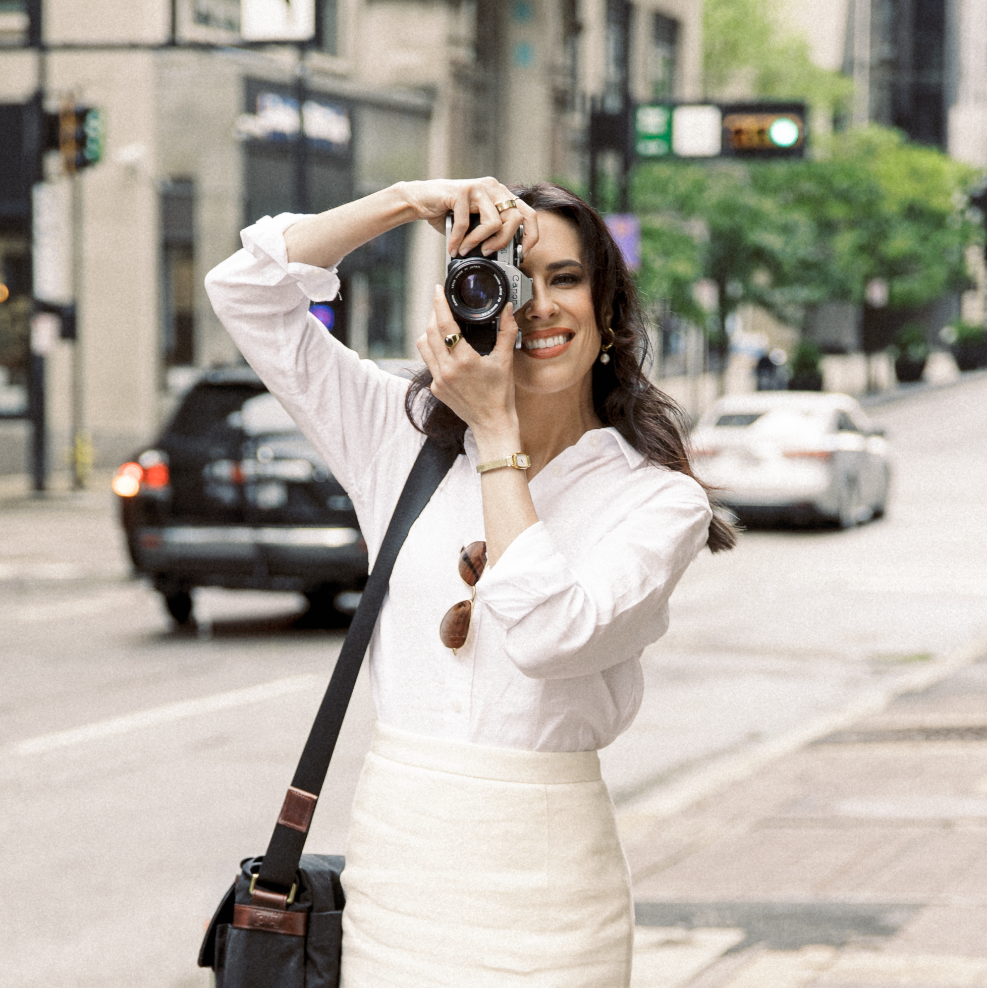 A woman standing on a city street, holding a camera up to her eye, smiling. She is wearing a white blouse and a cream skirt, with sunglasses hanging from her blouse, and has a black bag over her shoulder.