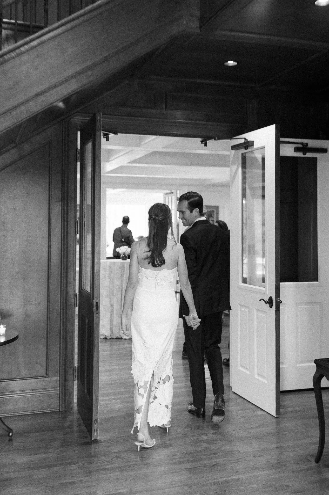 A couple holding hands and walking through a door at a wedding reception, with guests and a table in the background.