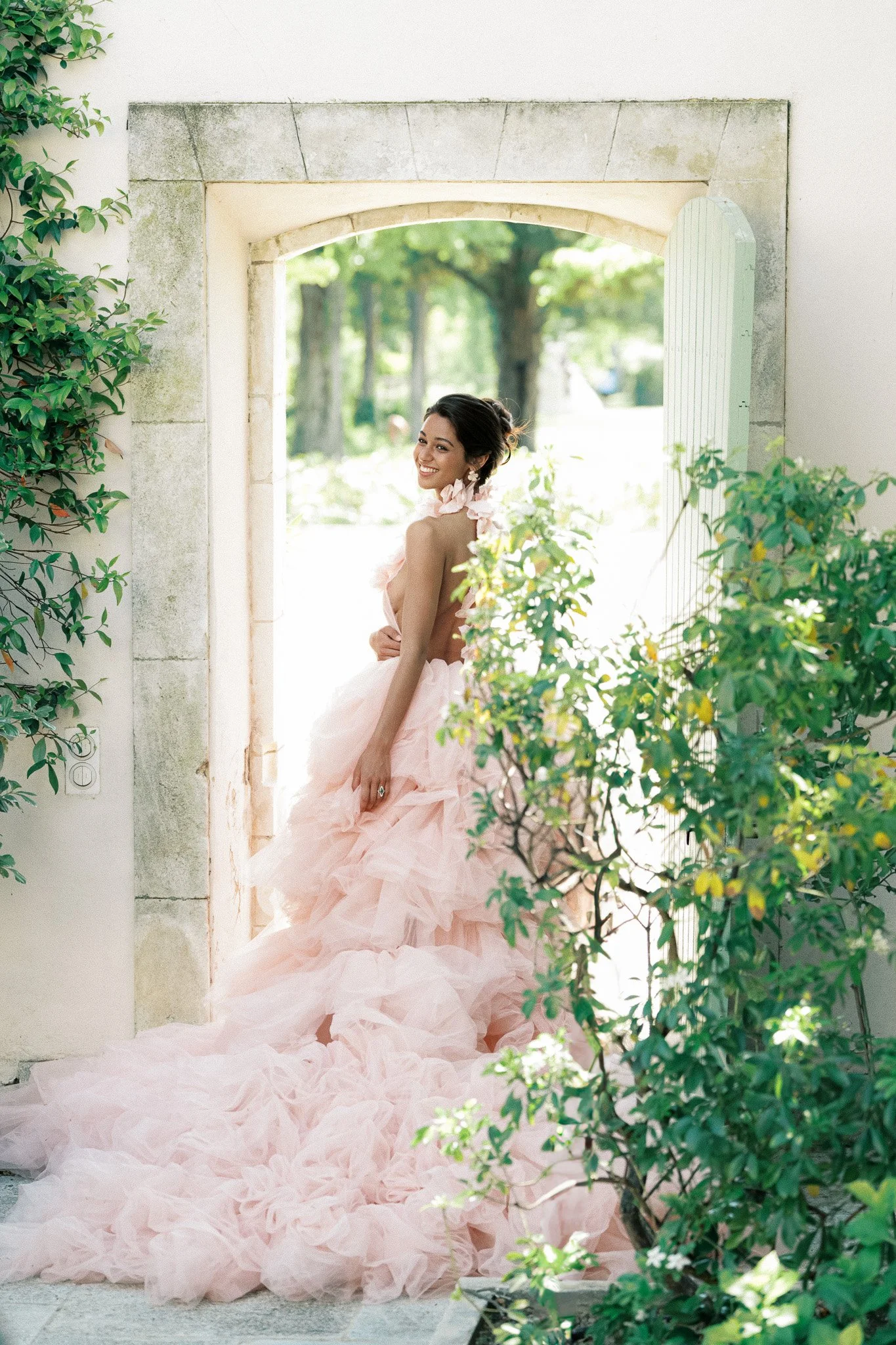 Smiling woman in a pink, ruffled gown standing in an open arched doorway surrounded by green foliage.