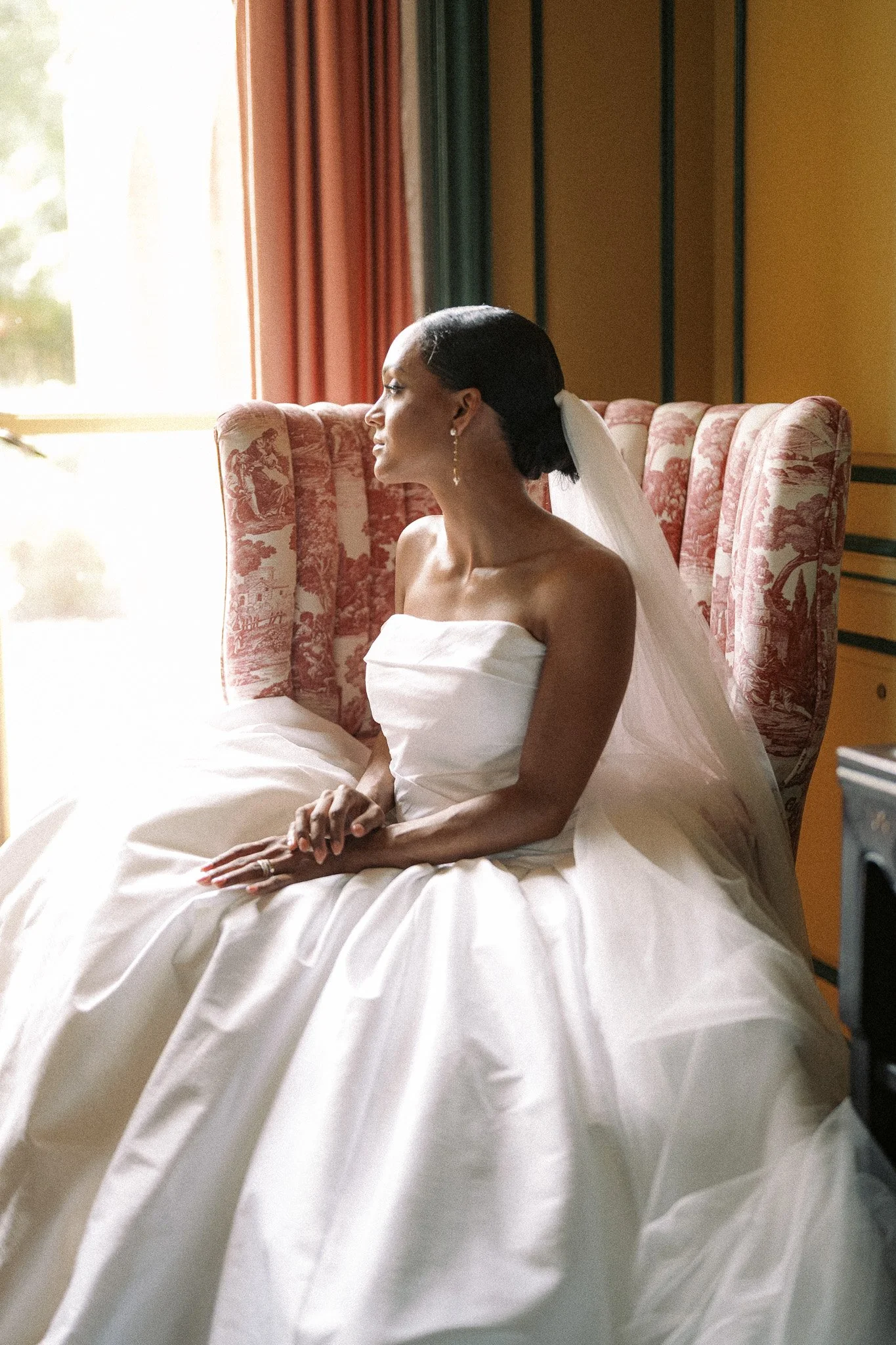 A woman in a wedding dress sitting on a bed near a window with curtains, illuminated by natural light.