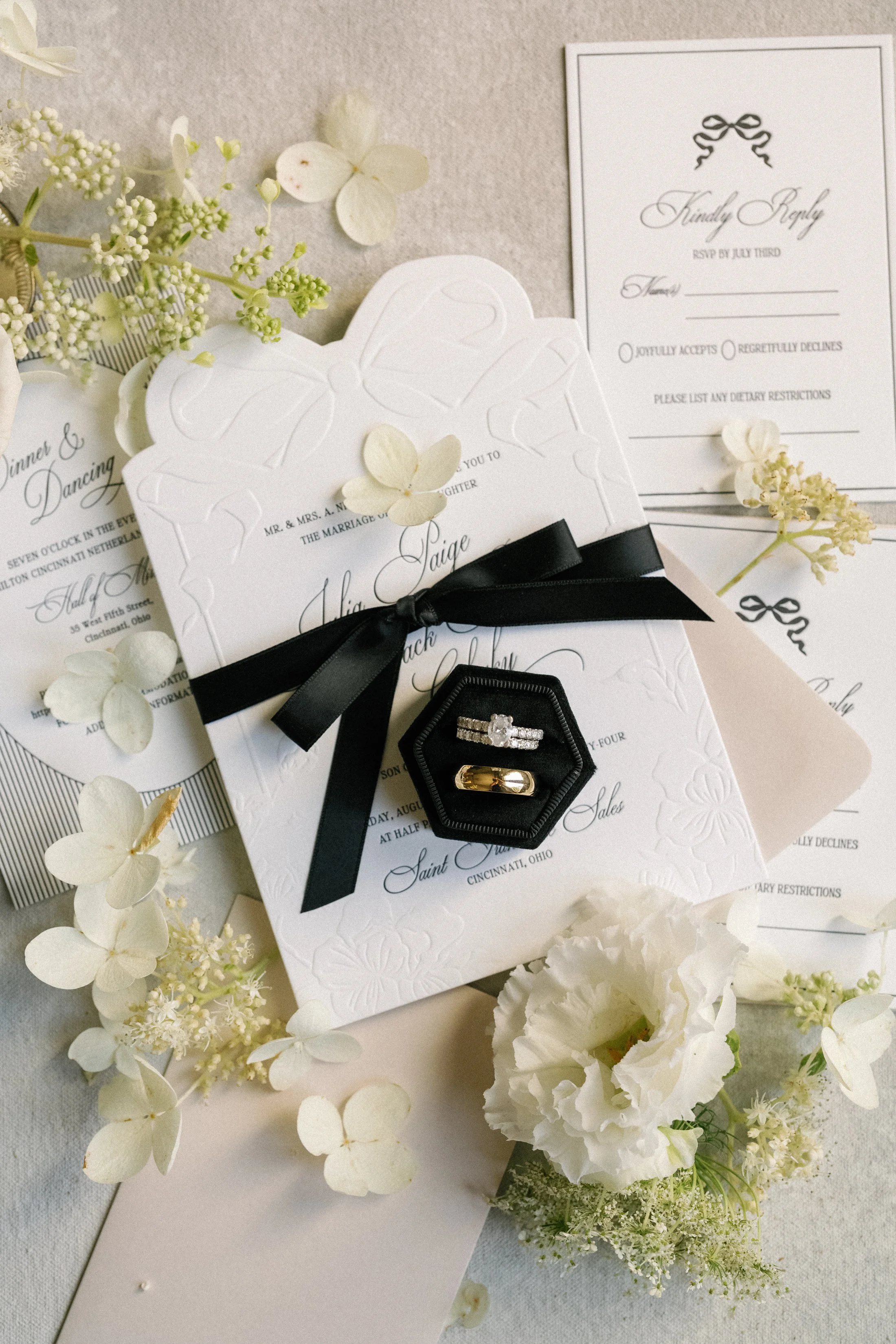 Wedding invitation set with black ribbon, floral decorations, and two rings in a black box.