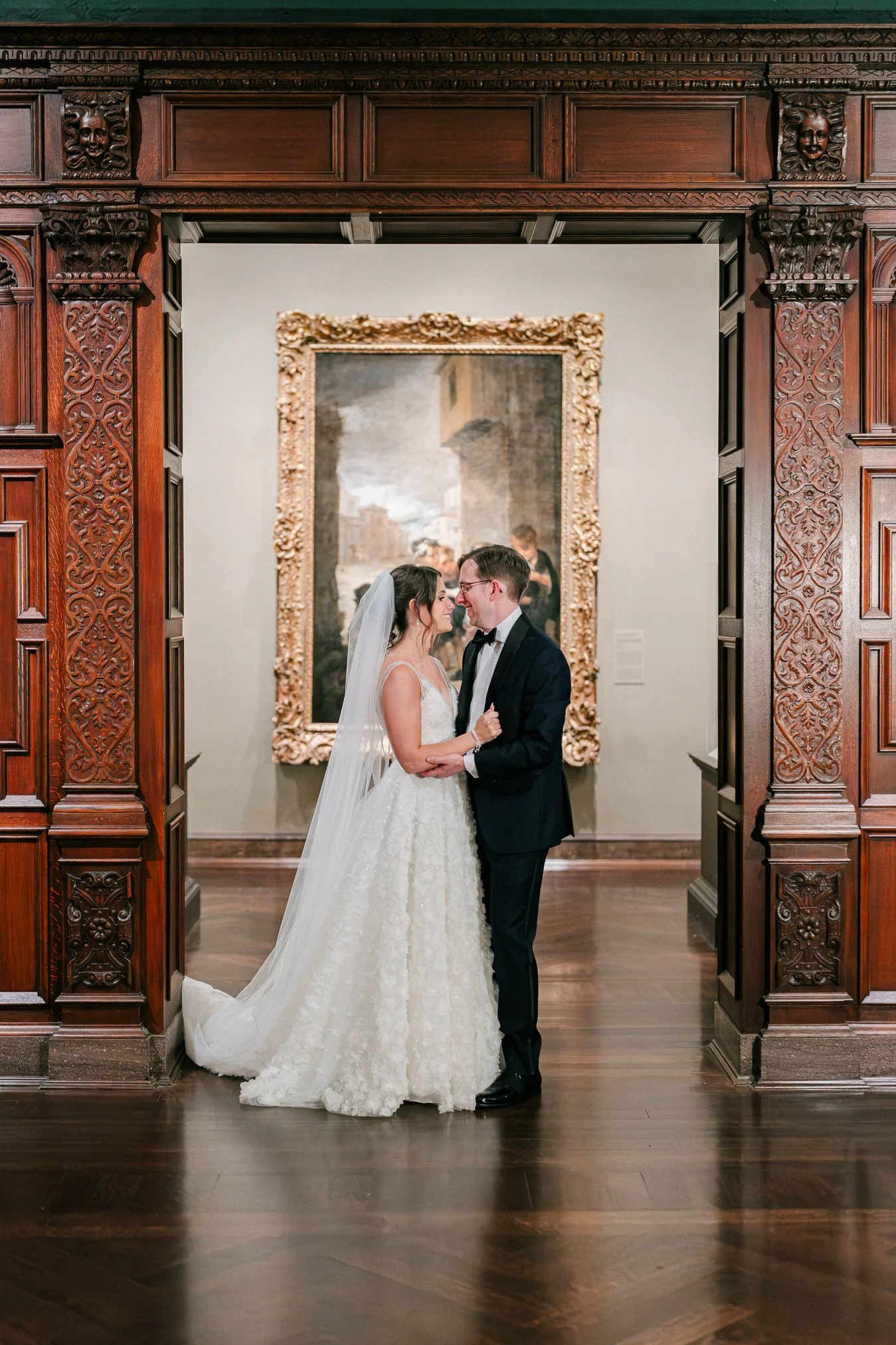 A bride and groom stand close together, smiling and touching noses, in an art gallery or museum with ornate wood paneling and a large framed painting in the background.