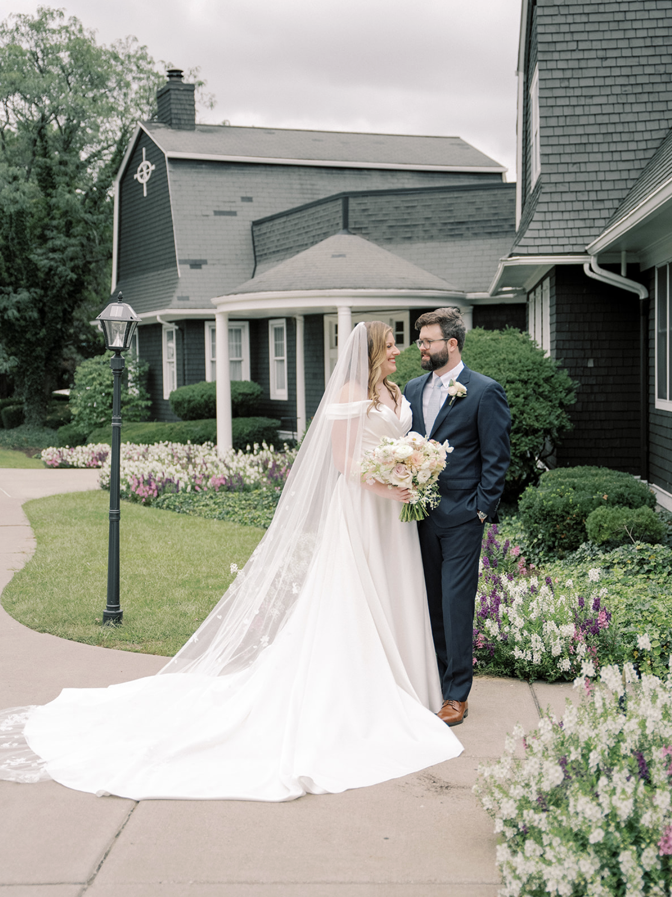 Bride and groom standing outside a house, gazing at each other. The bride wears a white wedding dress with a long veil and holds a bouquet of flowers. The groom is dressed in a navy suit with a light blue tie. The house has dark exterior shingles with white trim, and there are well-maintained bushes and flower beds around them.