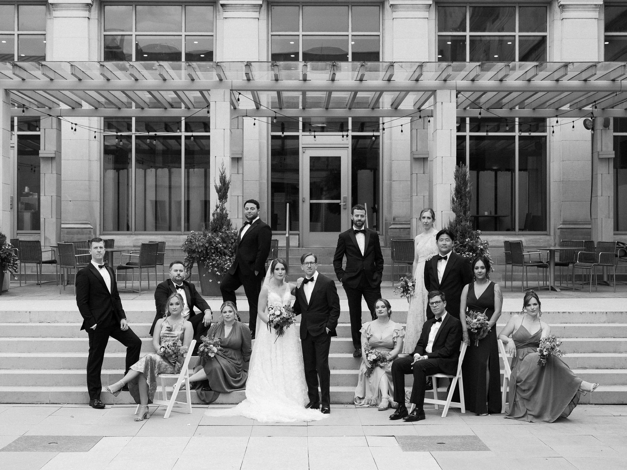 A wedding party posing on steps outside a building with large windows. The group includes men in tuxedos and women in dresses, with some women holding bouquets, and the bride in a white gown with a veil.