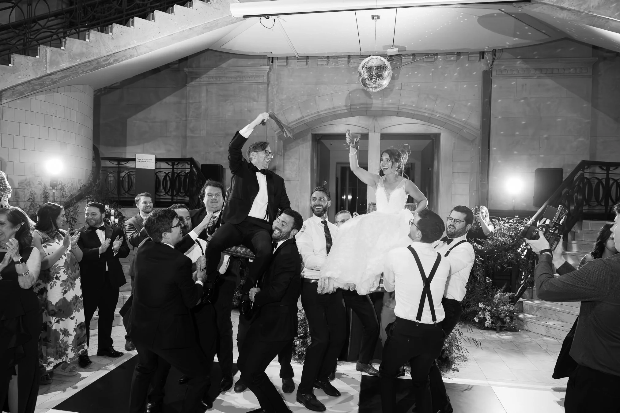 People celebrating at a wedding reception, lifting the bride and groom on chairs in a dance tradition, with a disco ball hanging from the ceiling, in a grand hall with stairs and floral decorations.
