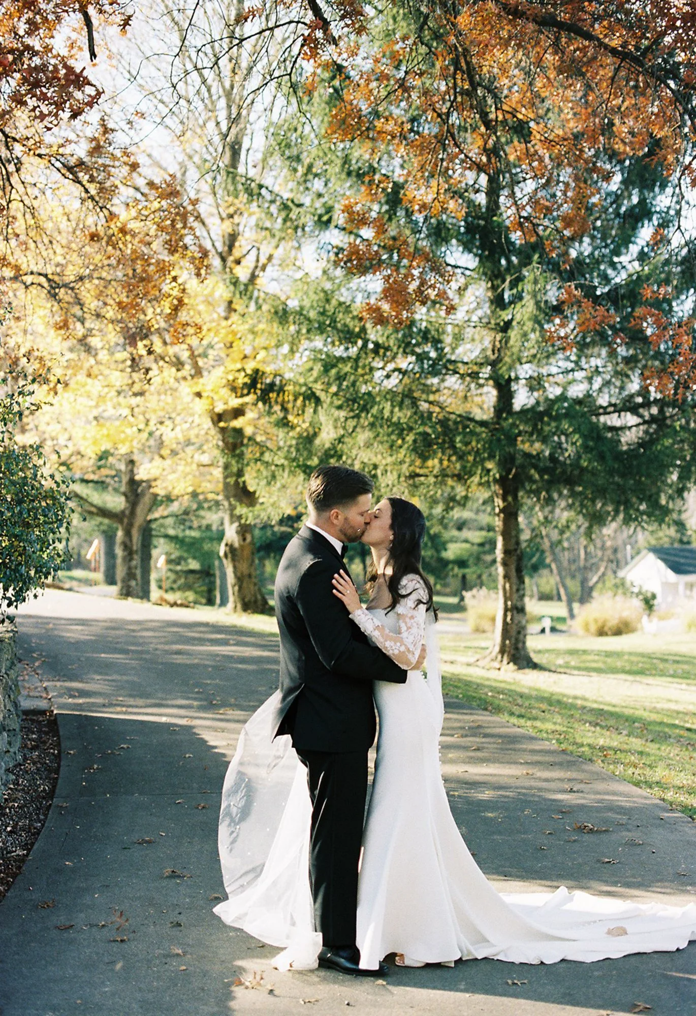 A couple in wedding attire sharing a kiss outdoors on a sunny day with autumn trees in the background.