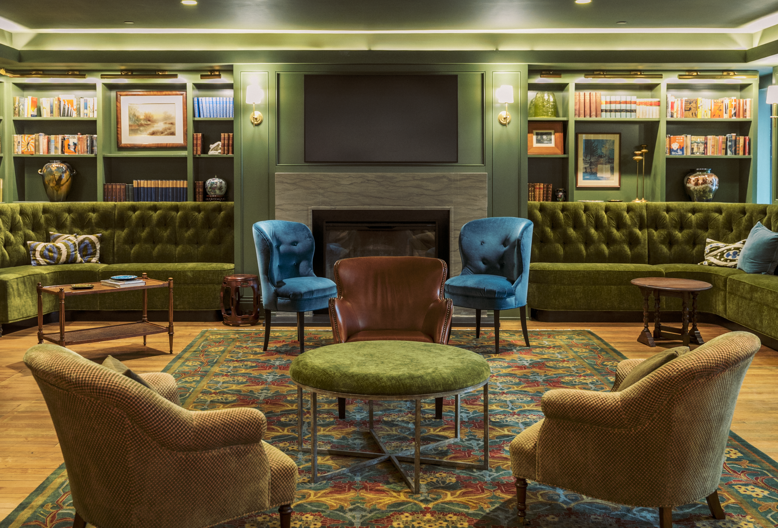 Living room with green walls, built-in bookshelves, and a fireplace. It has a colorful patterned rug, green and yellow armchairs, blue wingback chairs, a central green ottoman, a brown leather chair, and various side tables. A large flat-screen TV is mounted above the fireplace.