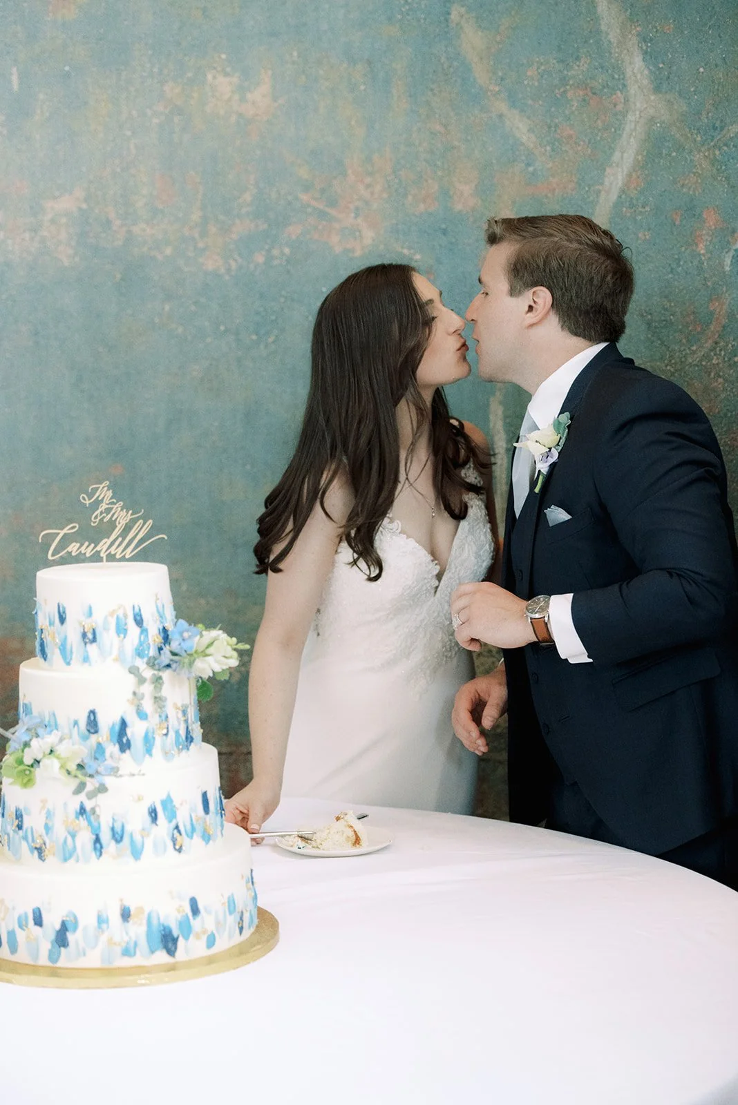 A bride and groom leaning in for a kiss at their wedding reception, with a three-tiered wedding cake on the table in front of them.