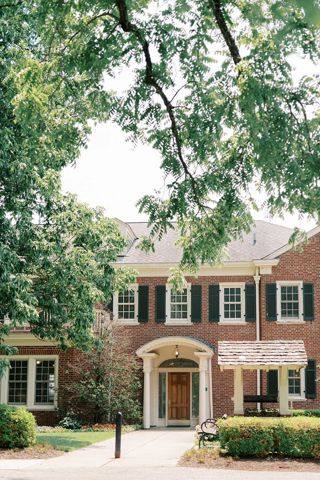 Front view of a brick house with multiple windows, black shutters, a wooden front door, and a small covered porch, surrounded by green trees and shrubs, with a sidewalk and bench in front.