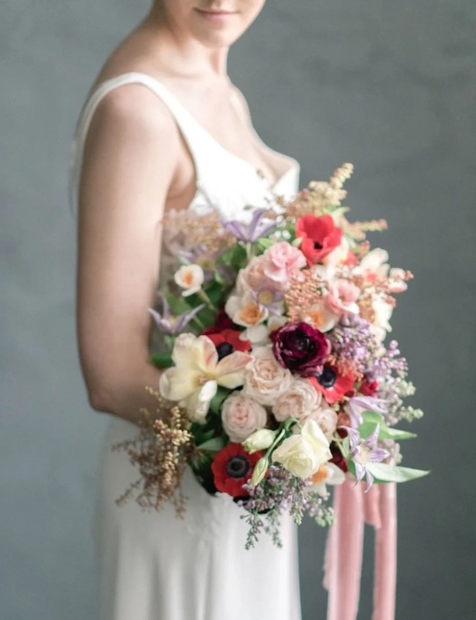 Bride holding a vibrant floral bouquet with roses and other mixed flowers in soft pink, red, and purple hues.