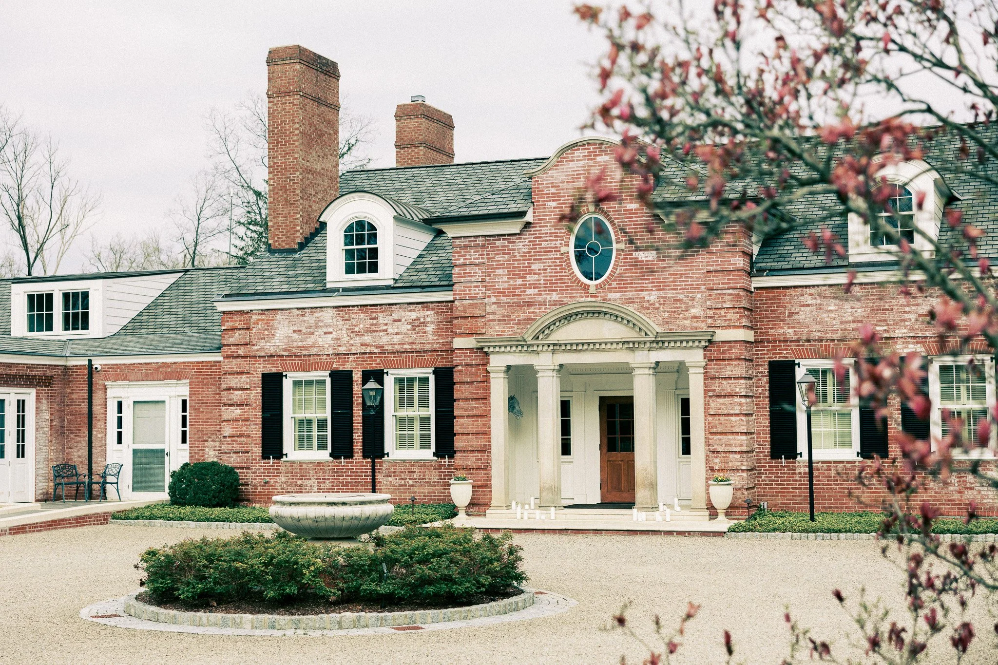 Front view of a large brick house with a circular driveway, garden bed, potted plants, and flowering tree branches in the foreground.