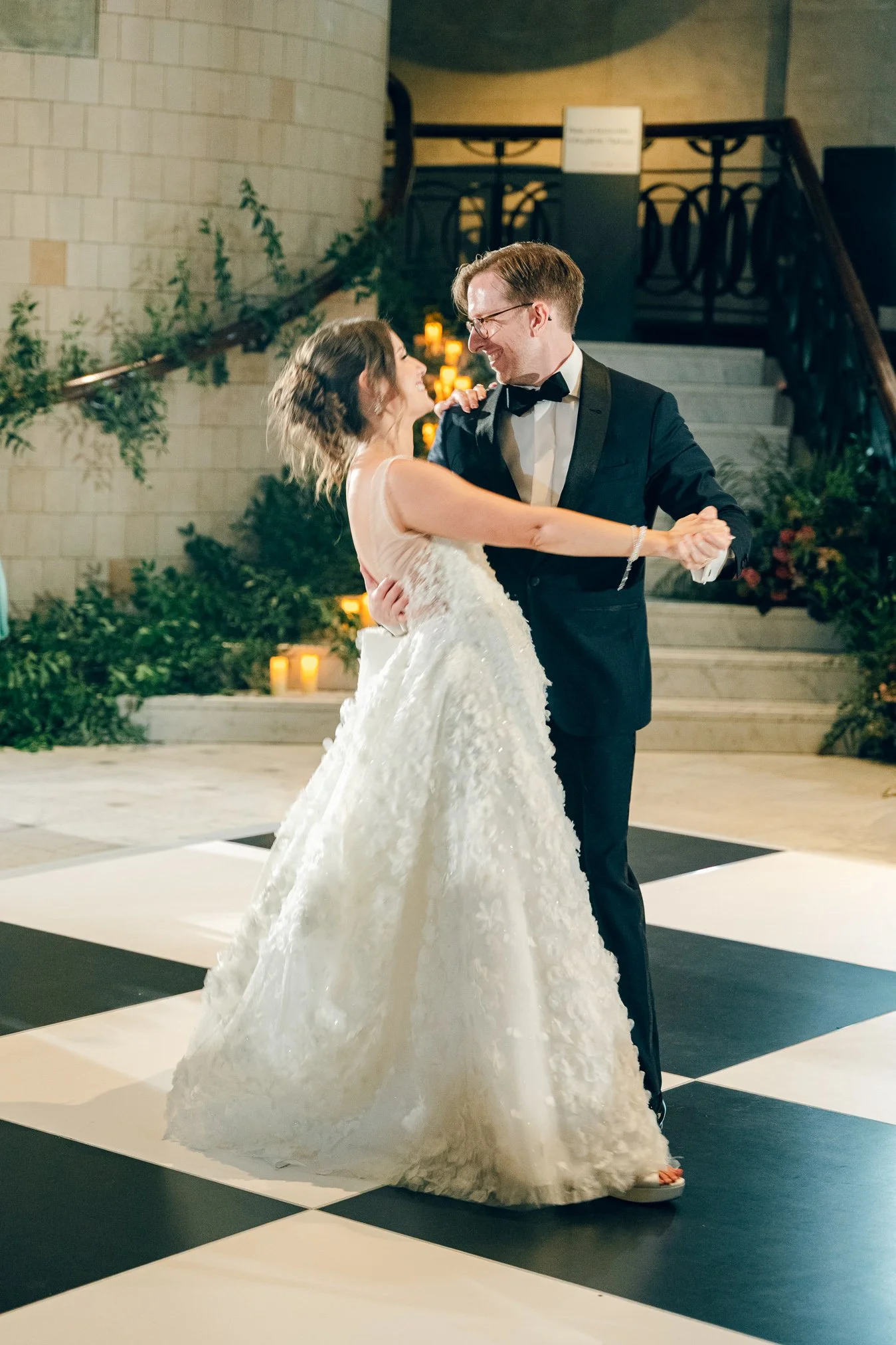 A bride and groom dancing together at their wedding reception indoors, with candles and greenery in the background.