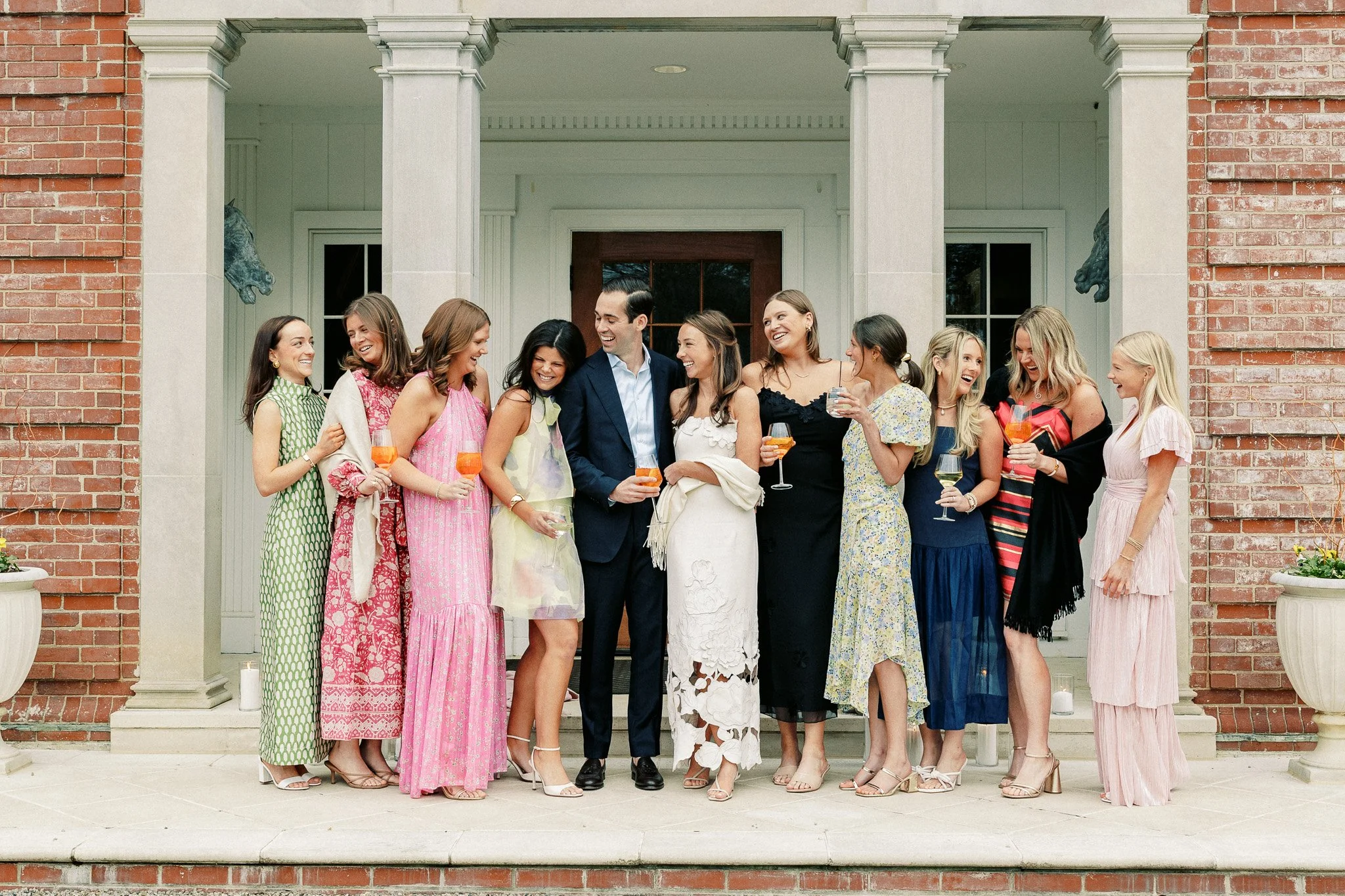 A group of fourteen women and one man standing on the front porch of a brick house, holding drinks and smiling. The women are wearing colorful dresses, and the man is dressed in a blue suit. They appear to be celebrating and enjoying each other's com