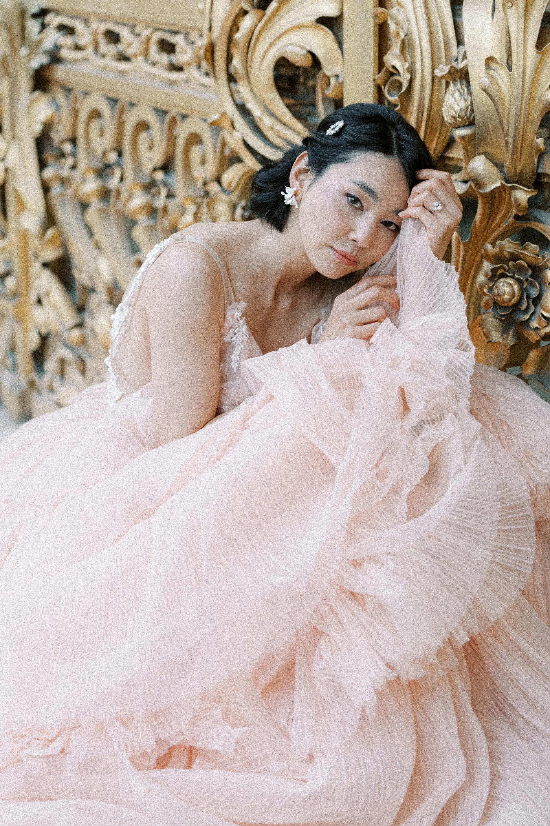 A woman in a pink pleated dress with floral embellishments, lying on a bed with a golden carved headboard, looking at the camera.