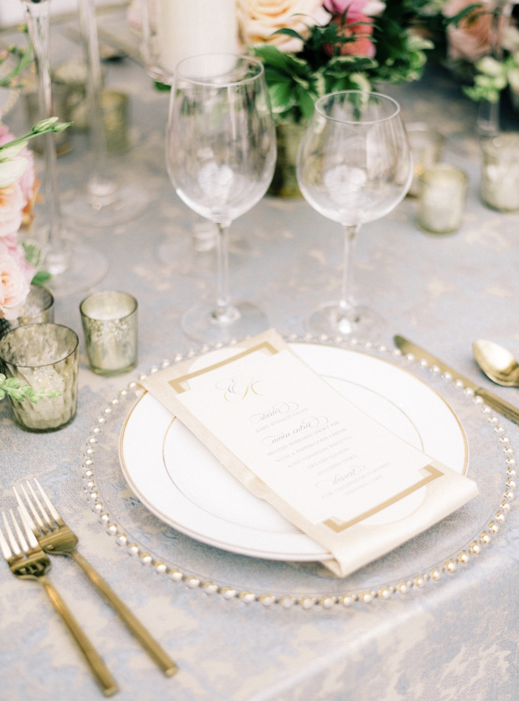 Elegant table setting with white and gold-rimmed plate, gold utensils, wine glasses, candles, floral arrangements, and a printed menu on a beige napkin.