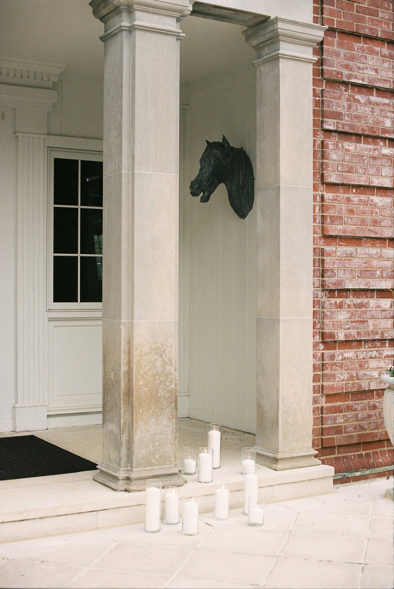 A black horse head sculpture mounted on a white wall inside an exterior brick and stone archway, with white candles at the base.