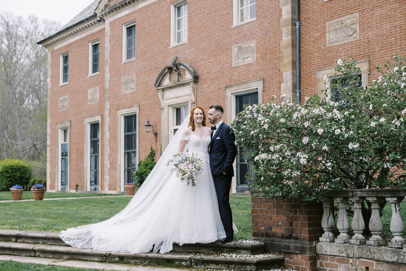 Bride and groom standing outside Peterloon Estate, with the bride in a white dress holding a bouquet and the groom in a black suit, surrounded by greenery. Cincinnati Wedding.