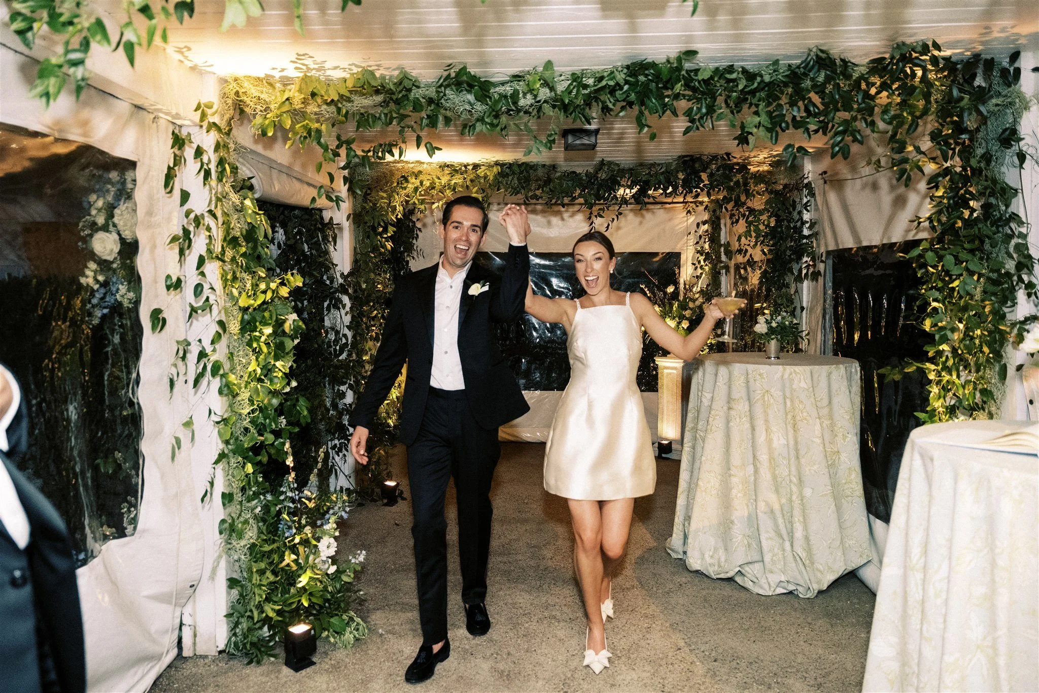 A newlywed couple celebrating indoors with greenery and floral decorations, with the groom and bride smiling and holding drinks.