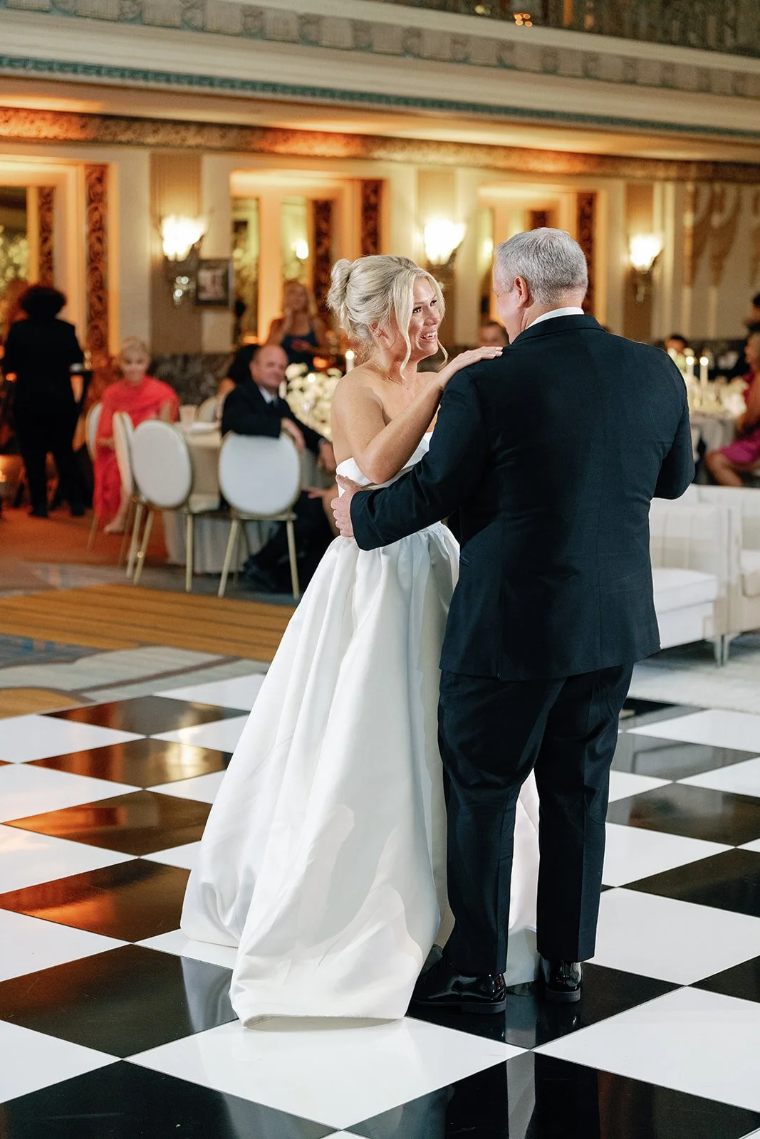 A bride and father sharing a dance at a wedding reception, with guests seated at decorated tables in the background.