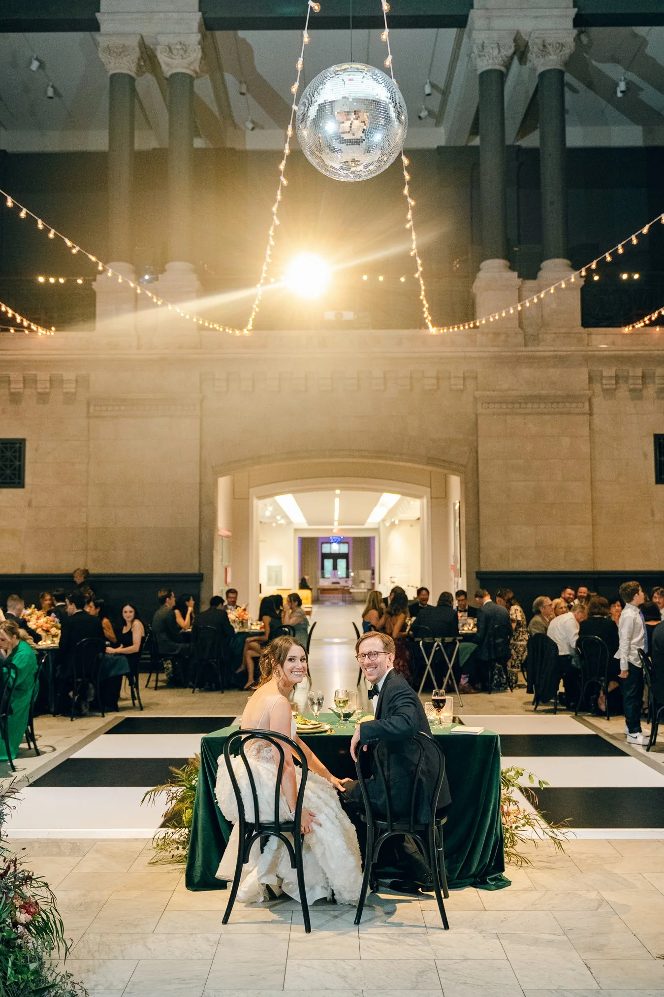 A wedding reception with the bride and groom sitting at a table, surrounded by guests, in a large elegant hall decorated with string lights and a disco ball.
