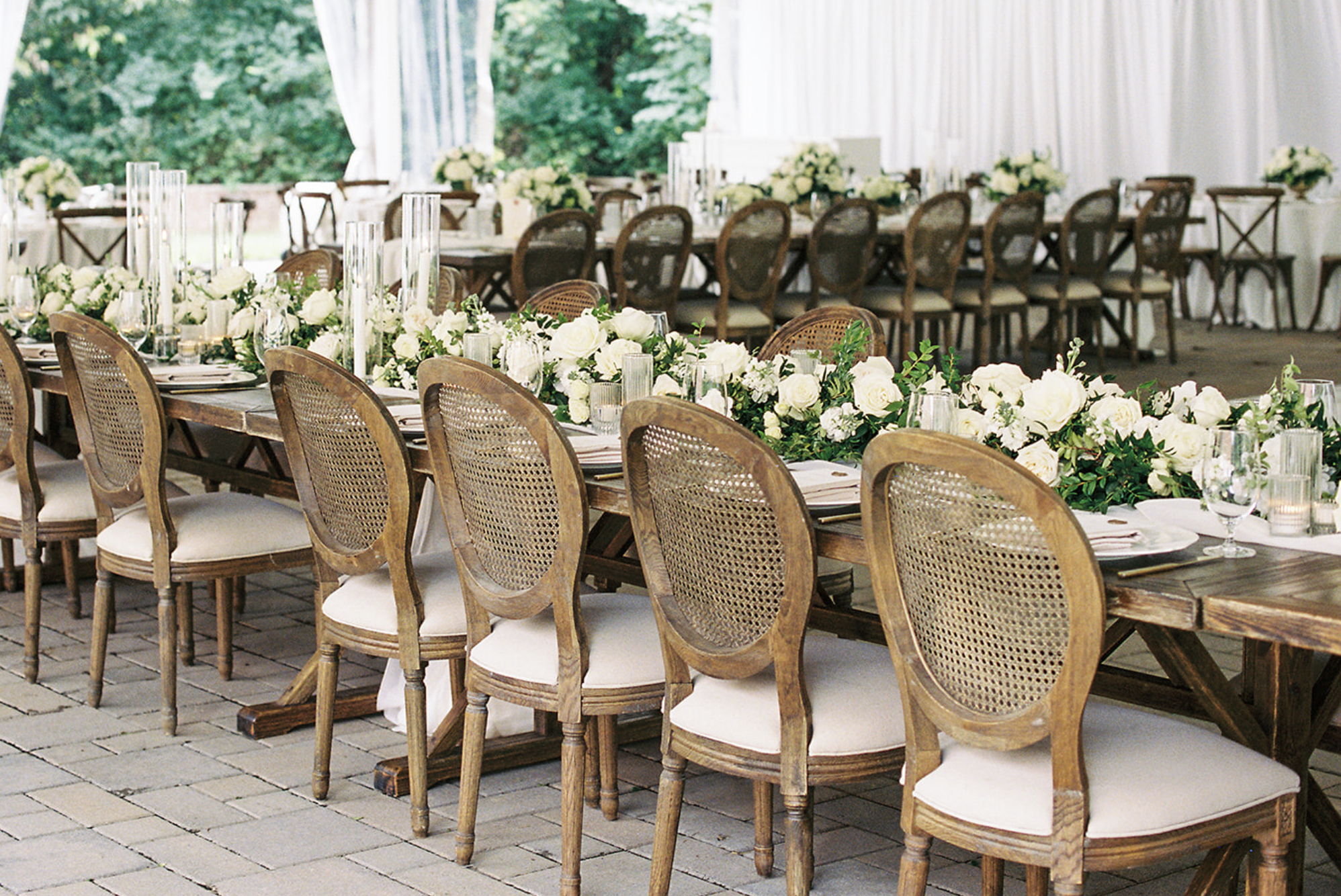 Elegant wedding reception setup with long wooden tables, cane-back chairs, white floral centerpieces, and candles, in a tented outdoor venue.