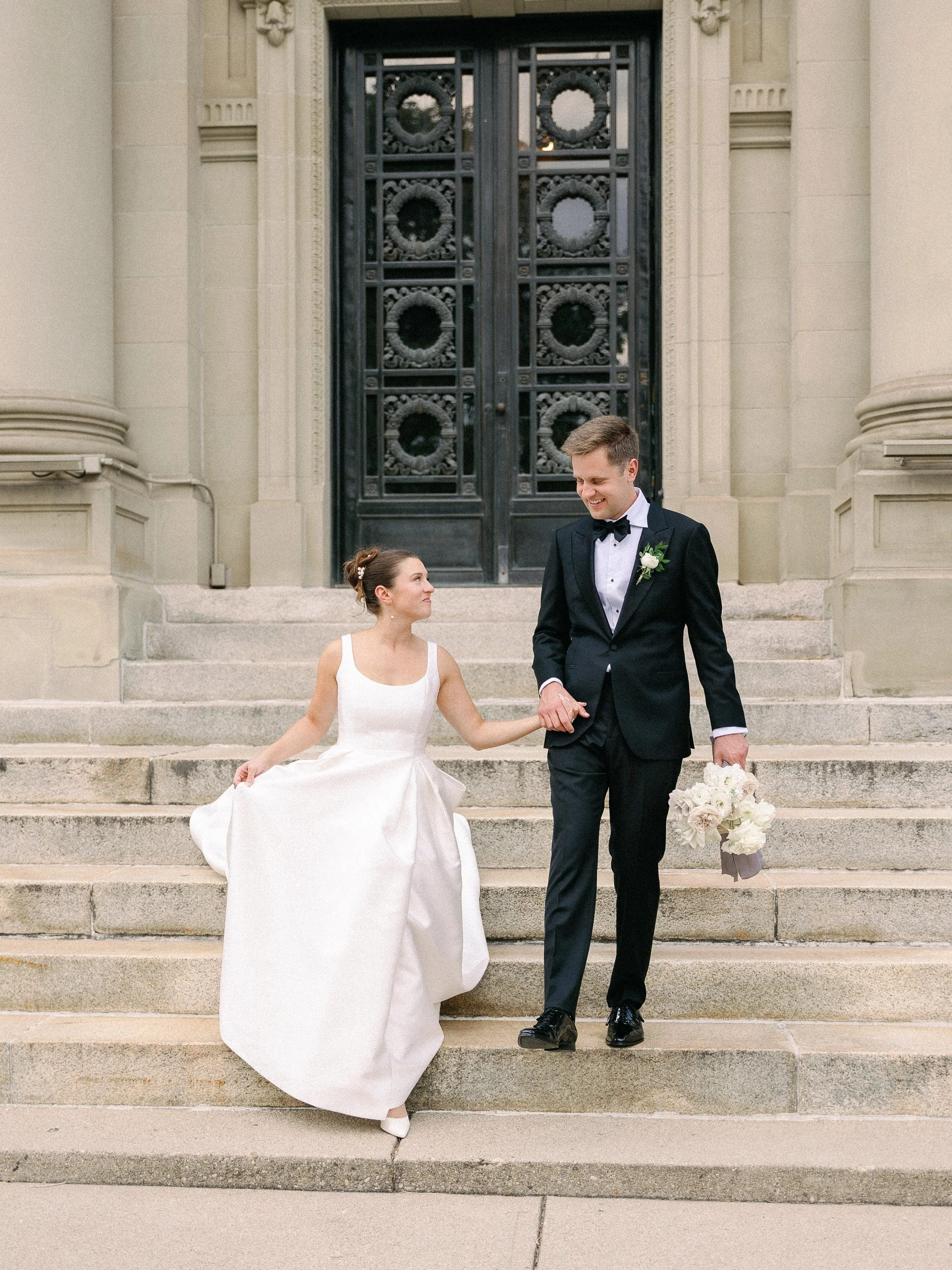 A bride and groom walking down the stairs outside a church, holding hands, with the bride wearing a white wedding dress and the groom in a black tuxedo holding a bouquet of flowers.