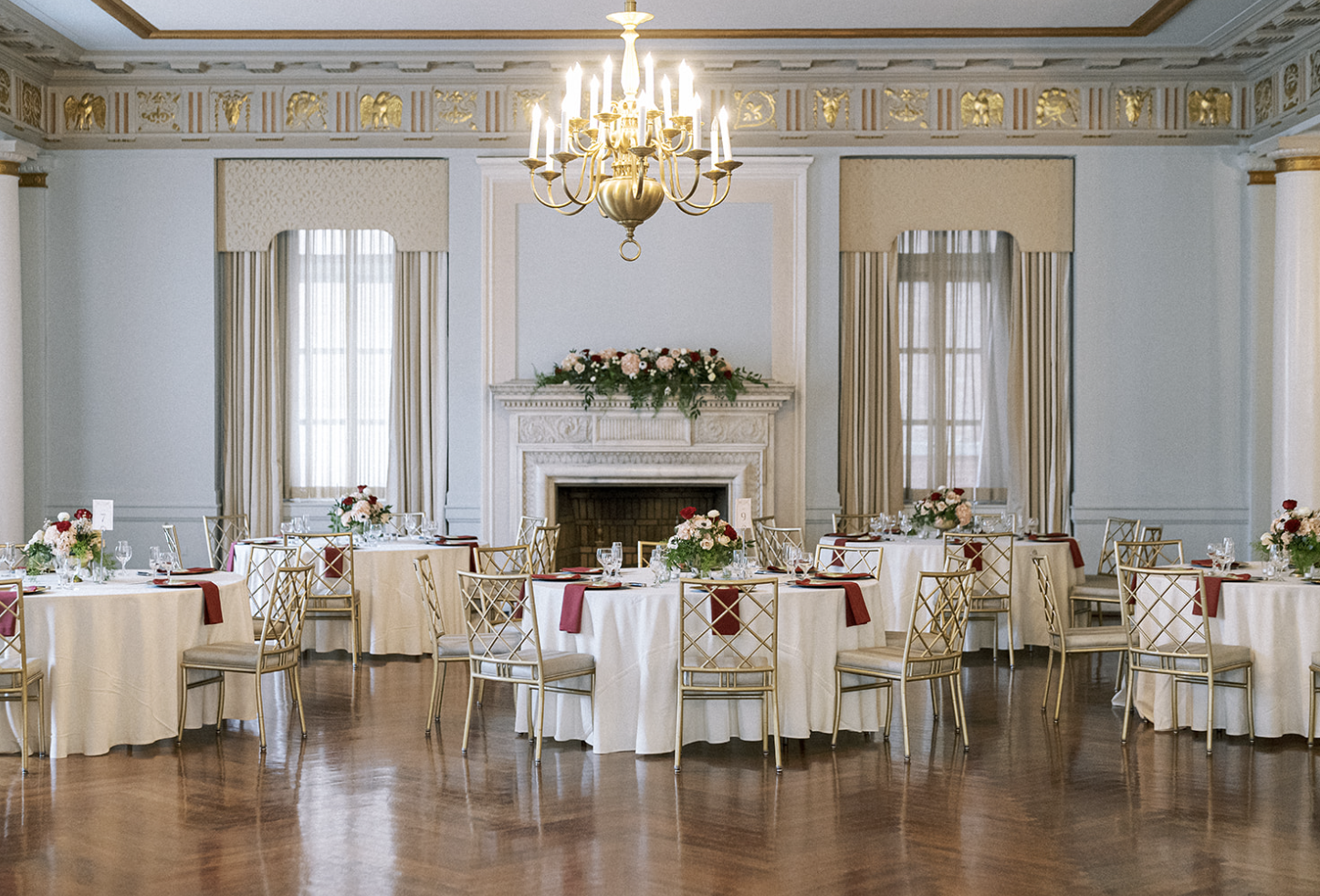 Elegant banquet hall with round tables set for an event, featuring white tablecloths, burgundy accents, gold chairs, a chandelier, and a decorative fireplace adorned with floral arrangements.