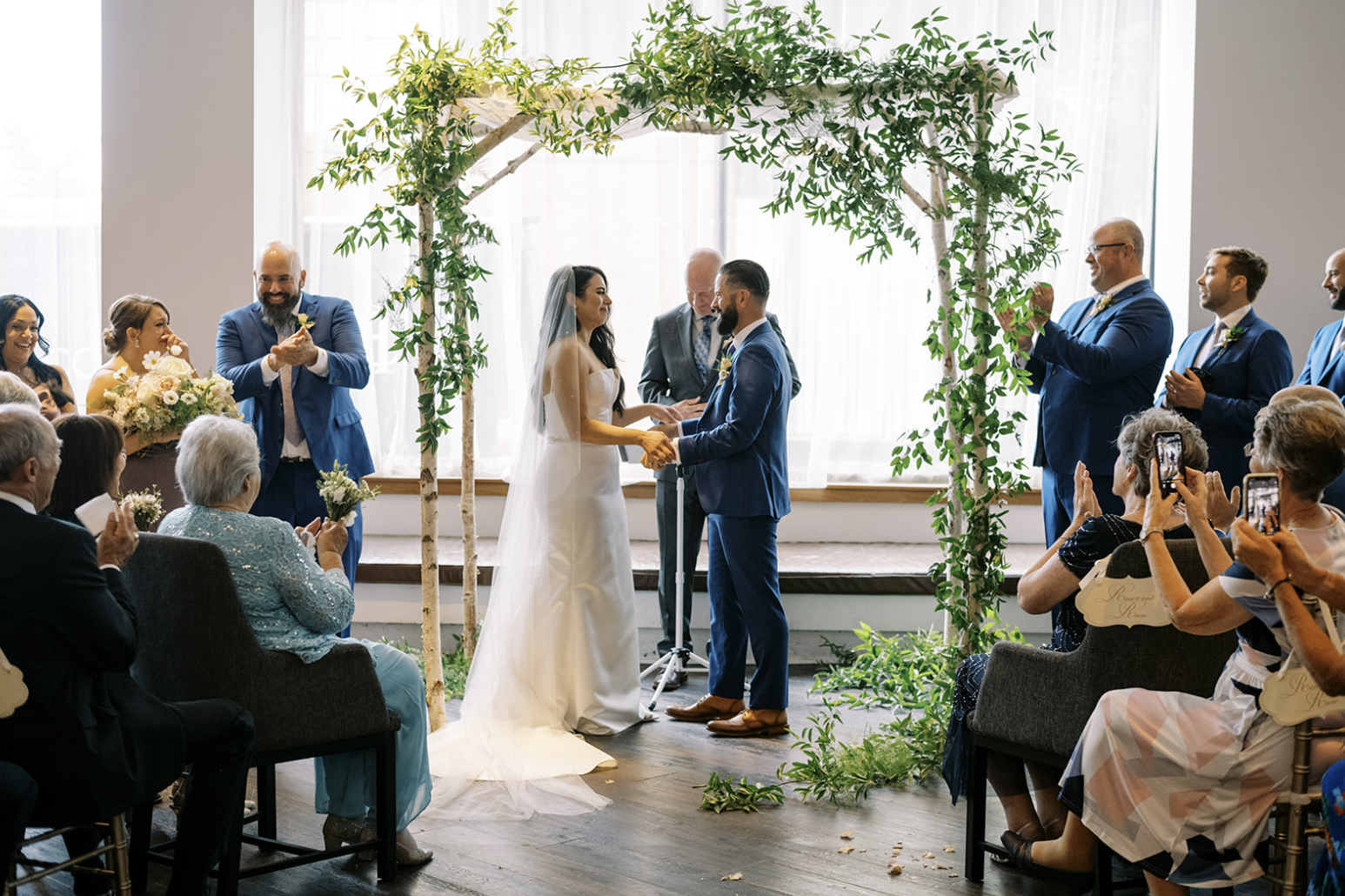A couple getting married under an arch with green foliage. The groom is wearing a blue suit, and the bride is in a white wedding dress and veil. They are holding hands, surrounded by wedding guests seated and clapping. The setting appears to be indoors.