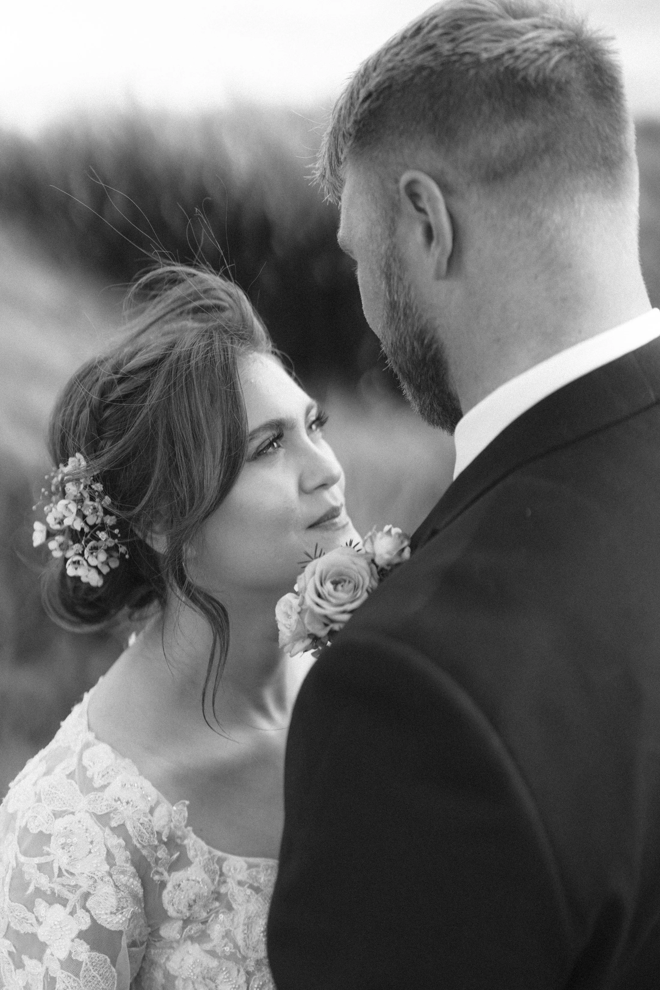 A black and white wedding photo of a bride and groom gazing into each other's eyes outdoors, with the bride holding a flower bouquet and wearing lace dress, and the groom in a suit.