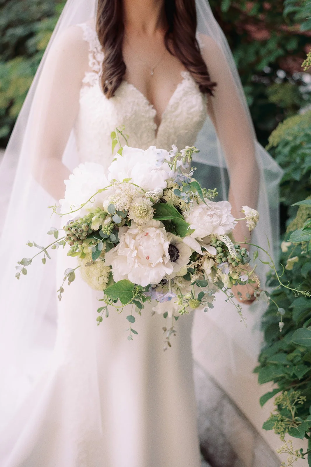 A bride in a white wedding gown holding a bouquet of white and pale purple flowers, standing outdoors with green foliage in the background.