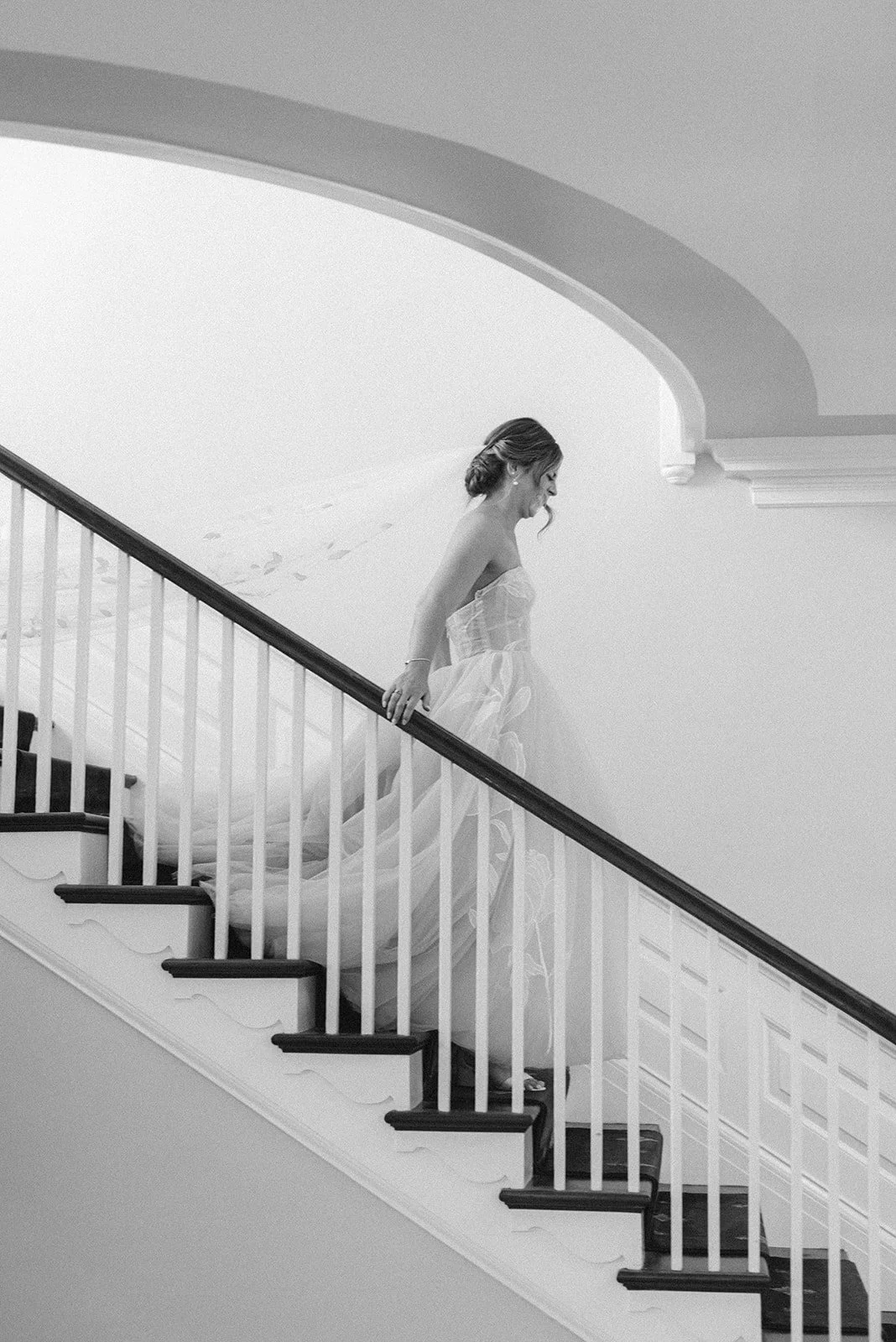 Black and white photo of a woman in a wedding dress walking down a staircase, smiling and looking down inside the French House in Cincinnati