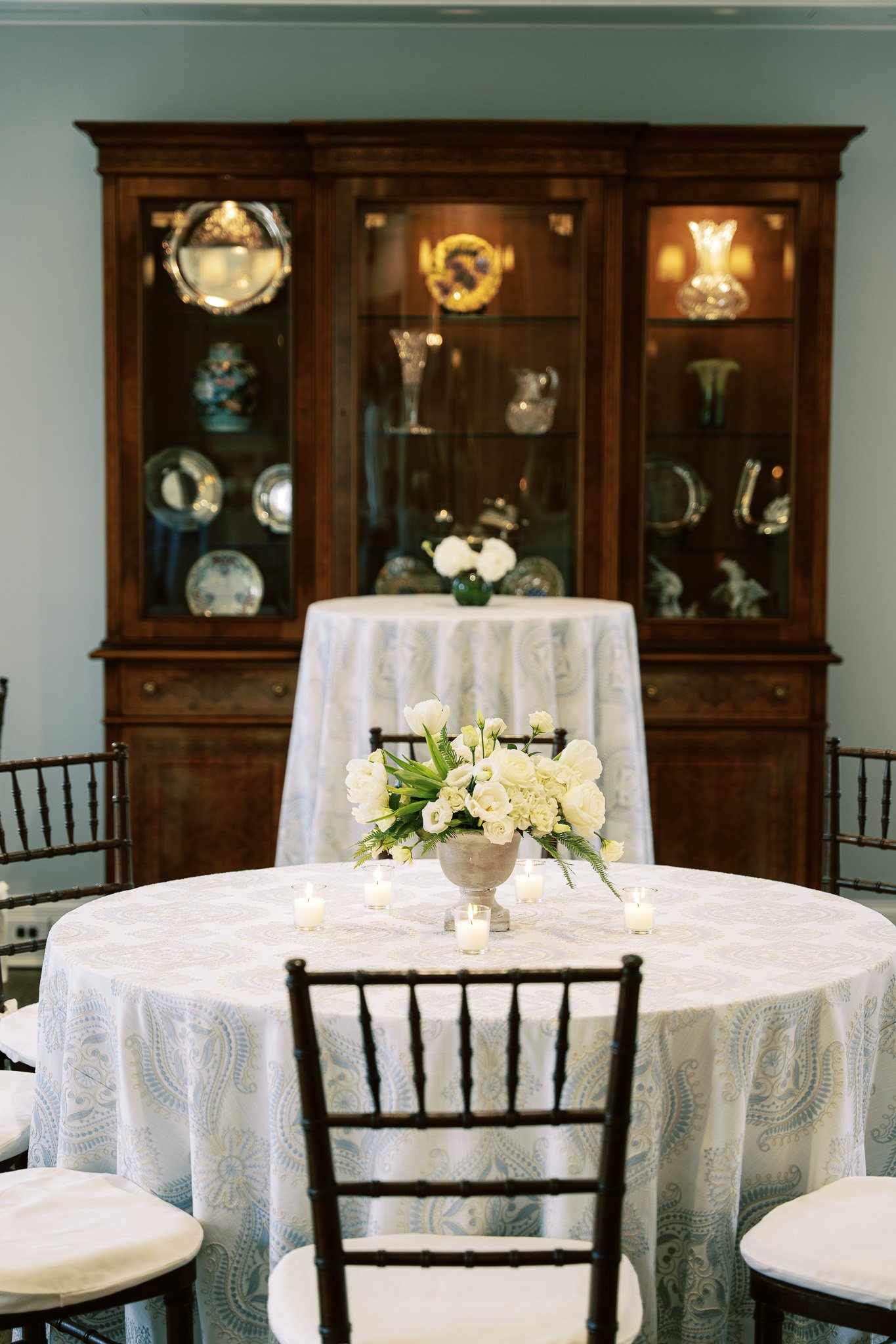 A dining room with a round table covered with a white tablecloth, a floral centerpiece with white flowers, small candles, a wooden hutch with glass doors displaying decorative glassware, and chairs with dark wood frames and white cushions.