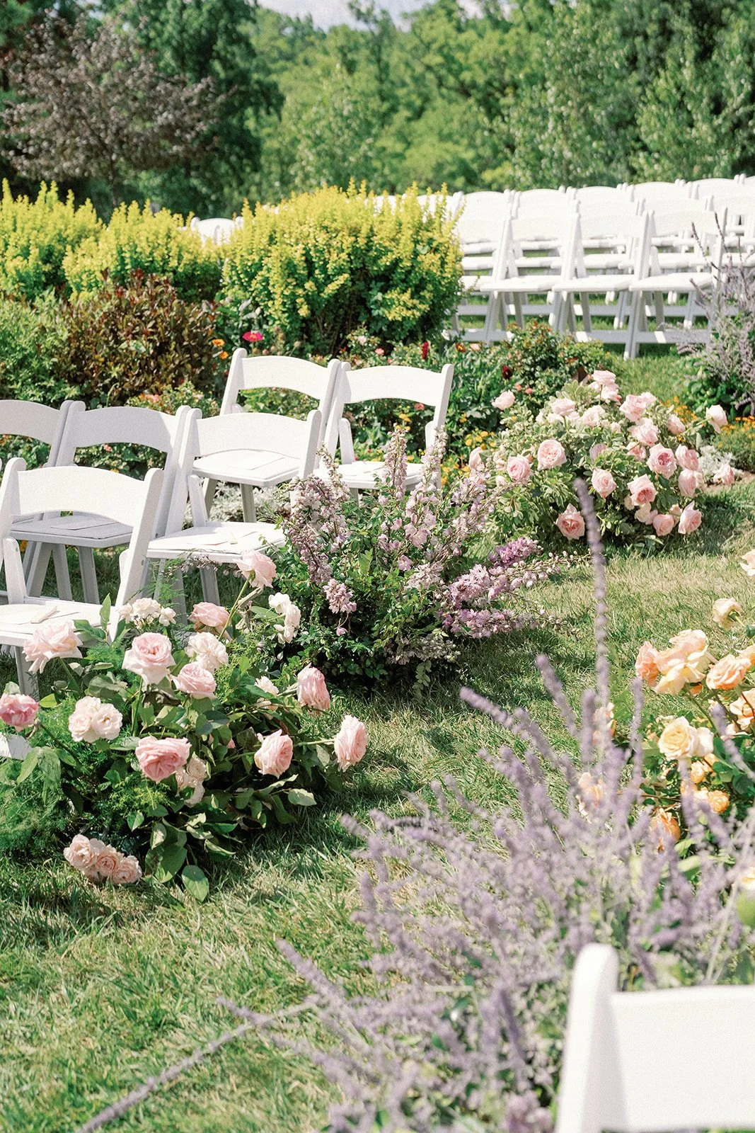White chairs arranged outdoors with pink and purple flowering bushes and greenery. A luxury garden wedding at French park in Cincinnati, Ohio