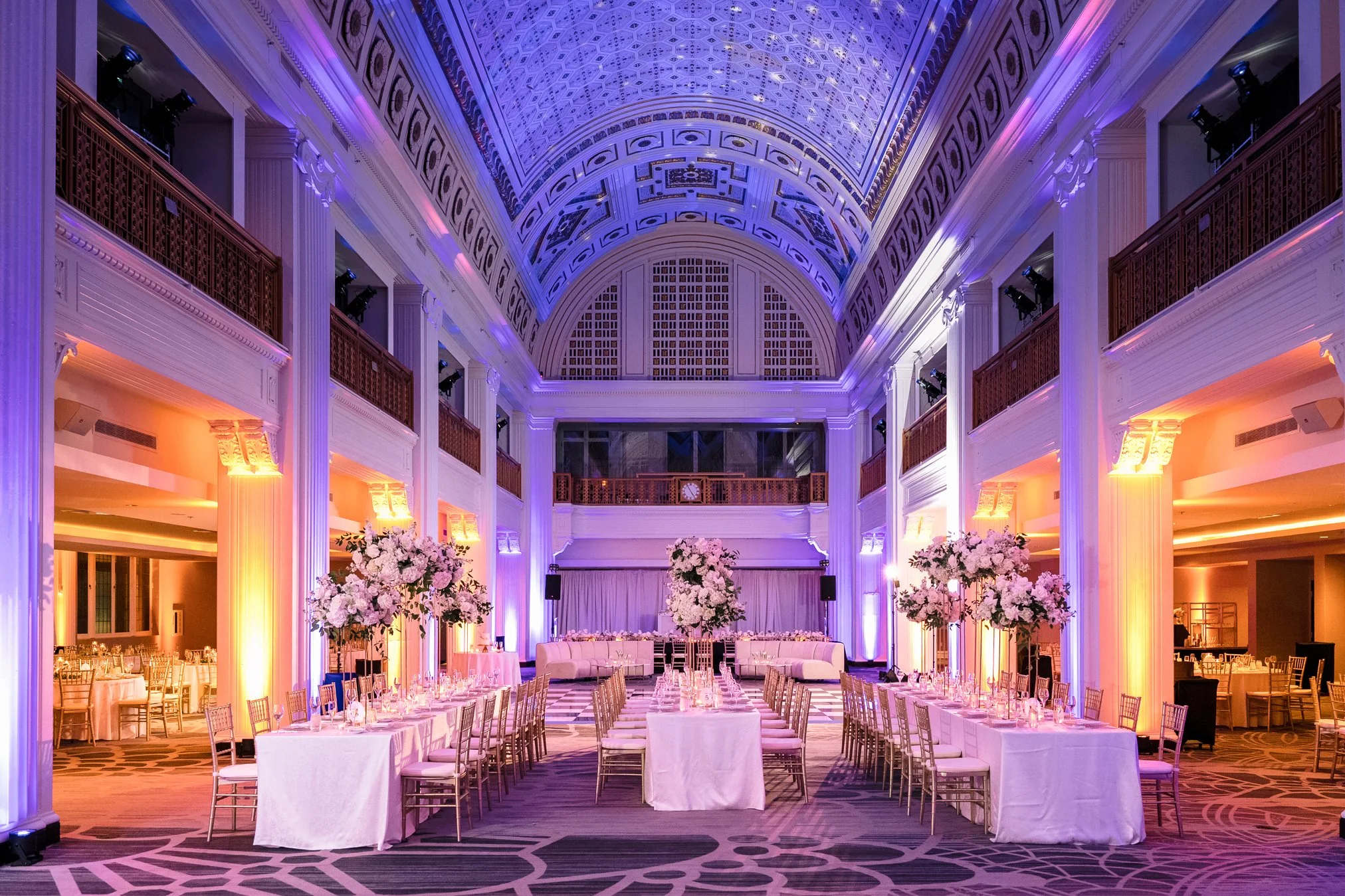 Elegant banquet hall with high arched ceilings, decorated with purple and warm lighting, featuring long tables with white tablecloths, tall floral centerpieces, and a stage at the far end with drapes.