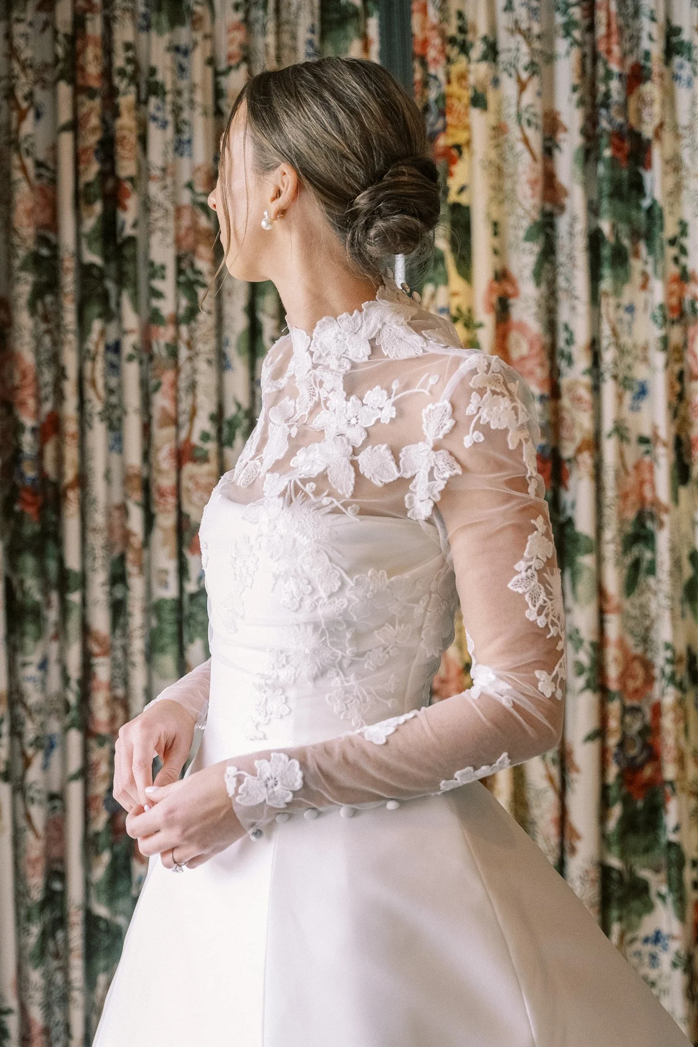 A woman wearing an elegant white wedding dress with lace floral details on the bodice and sleeves, standing in front of a floral curtain.