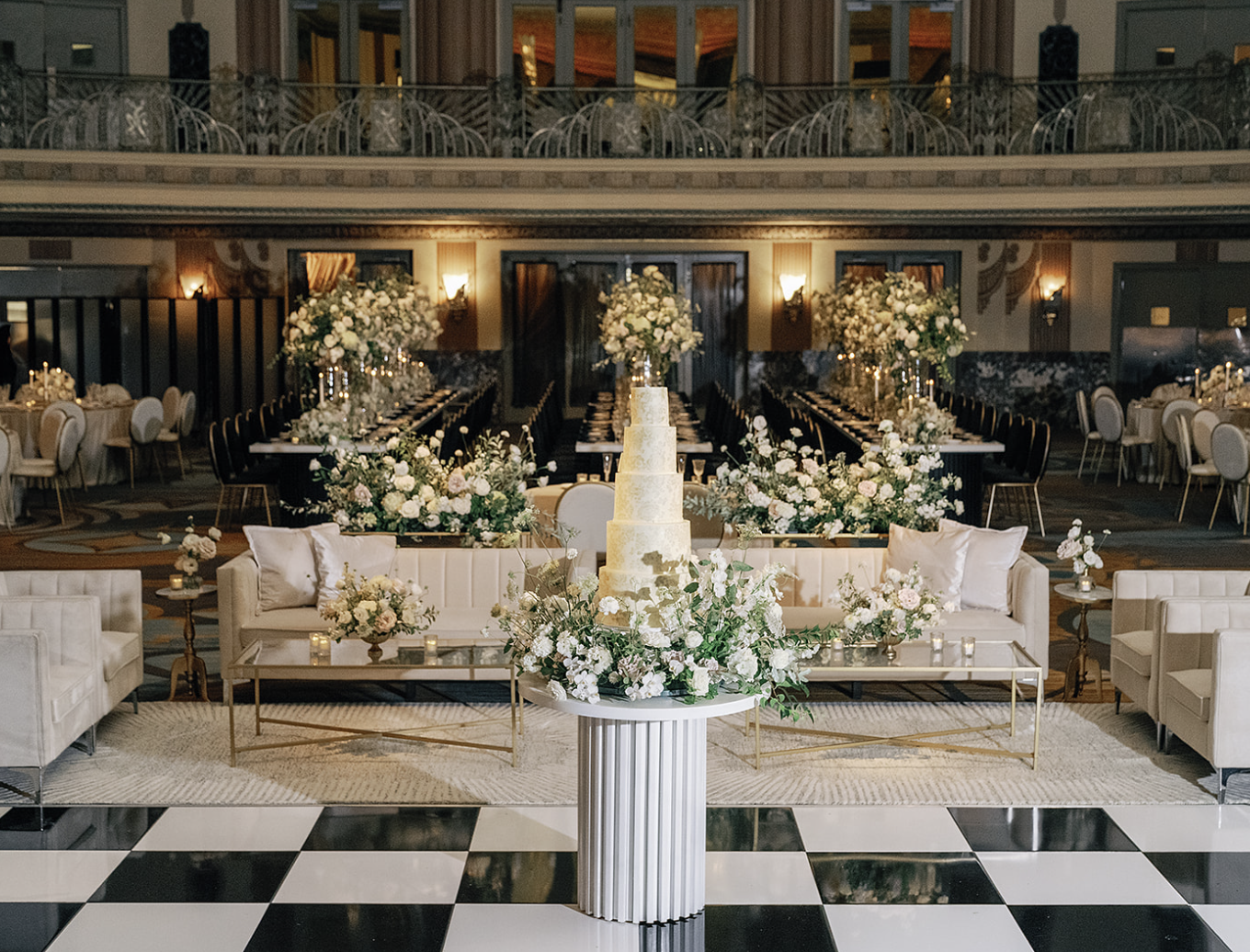 Elegant wedding reception setup with a tiered cake, floral arrangements, and seating area on a checkered floor.