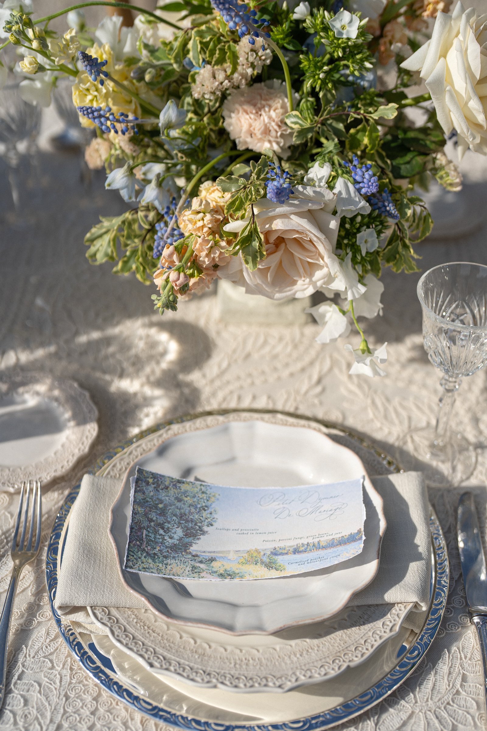 A tabletop setting with a floral centerpiece featuring white roses, blue muscari, pale pink carnations, and greenery. There are elegant white and blue china plates, a crystal glass, a menu card with a scenic illustration, and a white lace tablecloth.