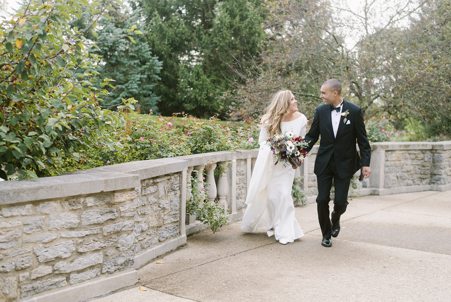 Bride and groom walking together in a garden, the bride in a white gown holding a bouquet, the groom in a black suit and bow tie.