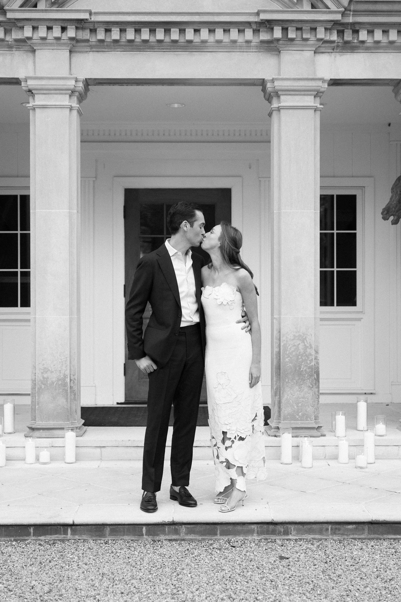 A black and white photo of a couple sharing a kiss on the front steps of a house, with candles surrounding them.