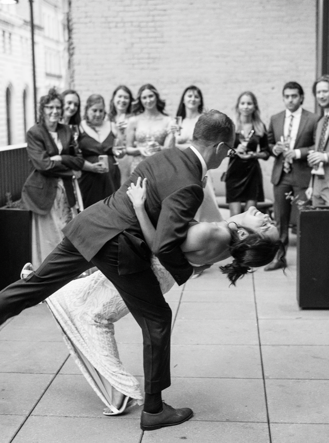 Couple dancing at a social gathering, with a group of people in the background holding drinks and watching. It appears to be a formal event.