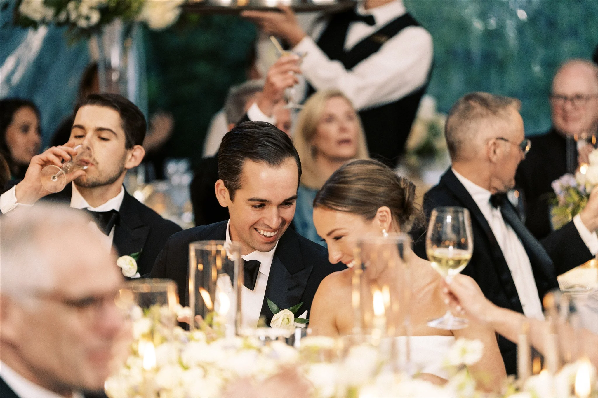 A wedding reception with a bride and groom smiling and talking, surrounded by guests in formal attire, some drinking wine and others in conversation, at a decorated table.