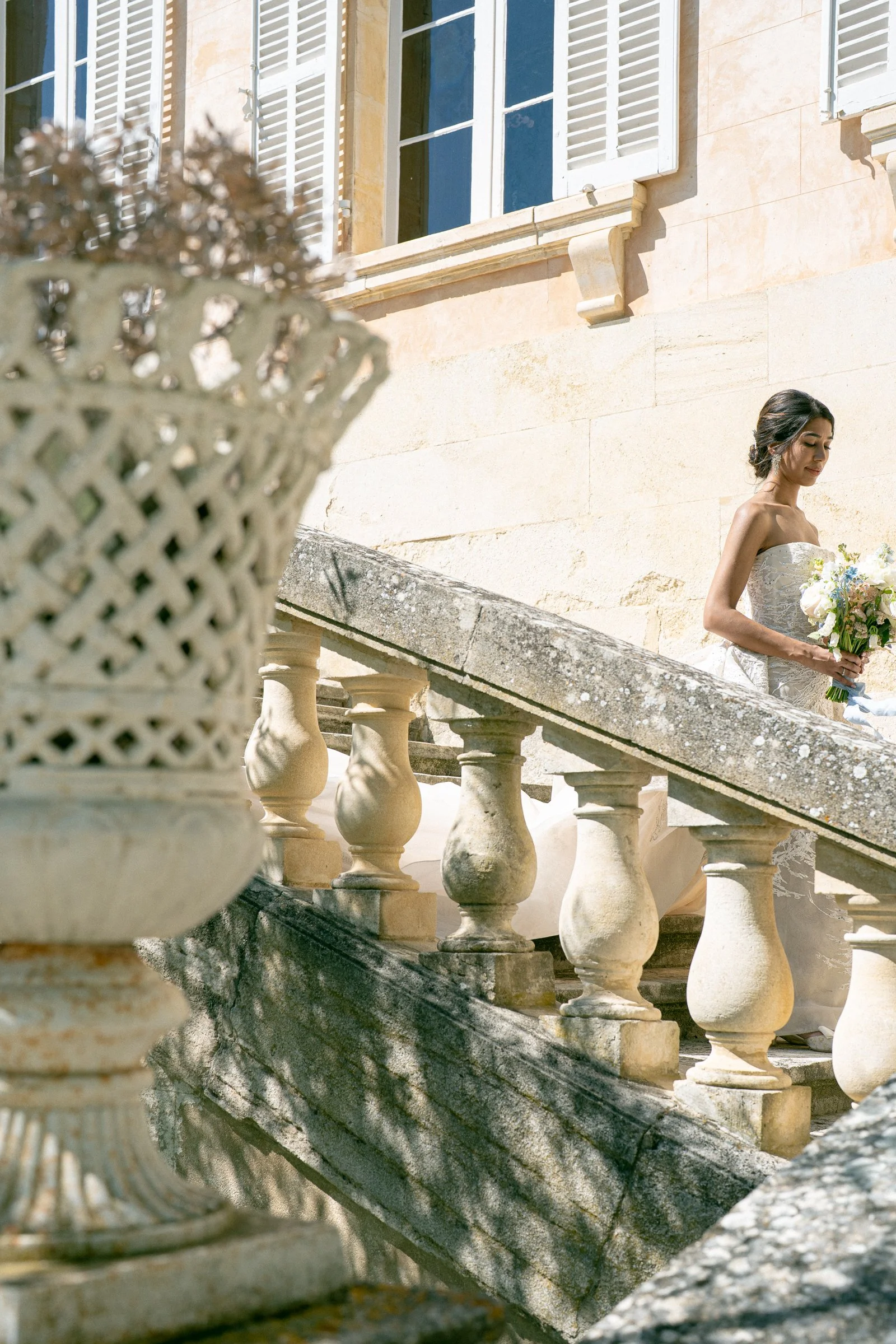 A woman in a wedding dress holding a bouquet standing on a stone staircase outside a building with beige stone walls and white window shutters, with a decorative urn in the foreground.